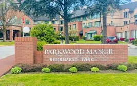 A brick entrance sign reads 'Parkwood Manor, Retirement Living,' in front of apartment buildings with trees and parked cars. Located in Coquitlam. 