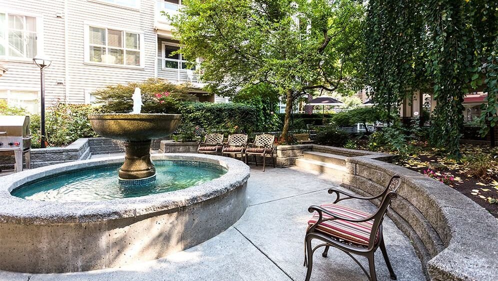 A concrete fountain with a round basin and a taller centerpiece in a courtyard garden. Surrounding the fountain are benches, trees, shrubs, and a building with multiple windows. Mayfair Terrace, retirement home. 
