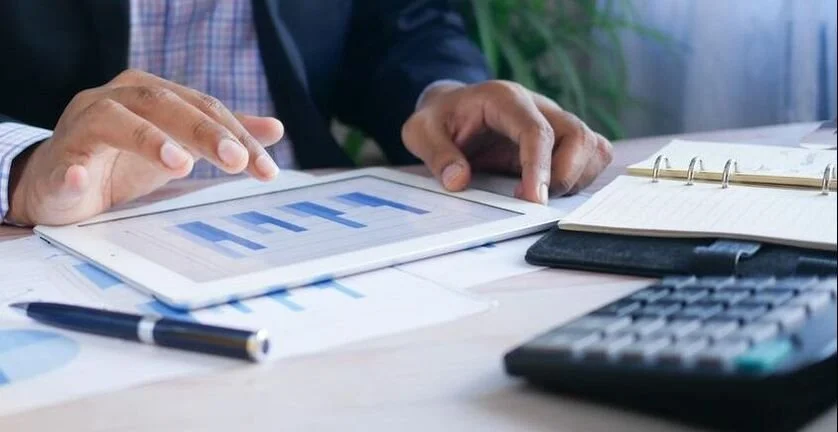 Hands of a person reviewing financial charts and data on a tablet, with a pen, notebook, and calculator on the desk.
