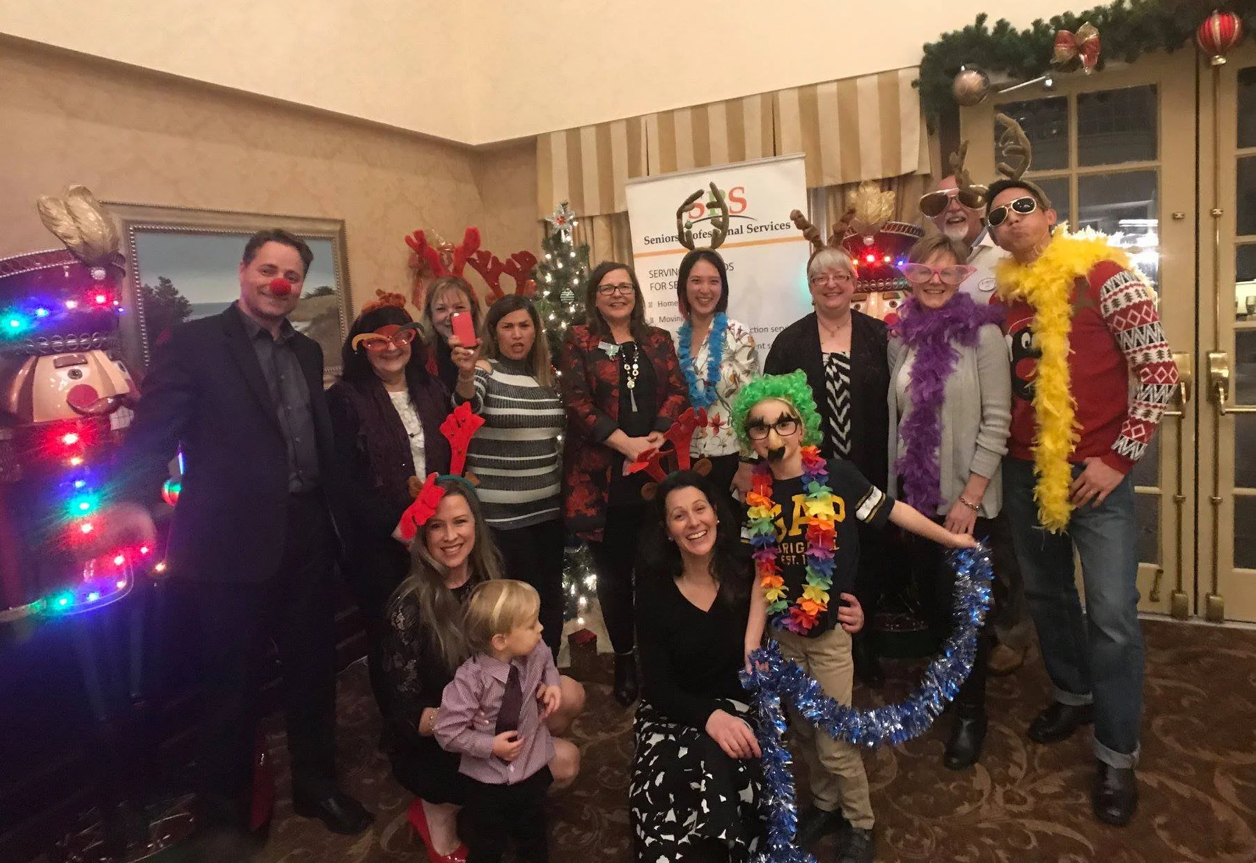 Group of people celebrating Christmas, dressed in festive costumes and accessories like reindeer antlers, standing in front of a decorated Christmas tree at a retirement home in Coquitlam.