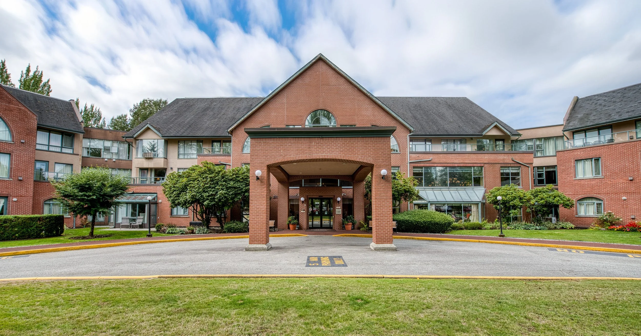 A brick entrance sign reads 'Parkwood Manor, Retirement Living,' in front of apartment buildings with trees and parked cars. Located in Coquitlam. 