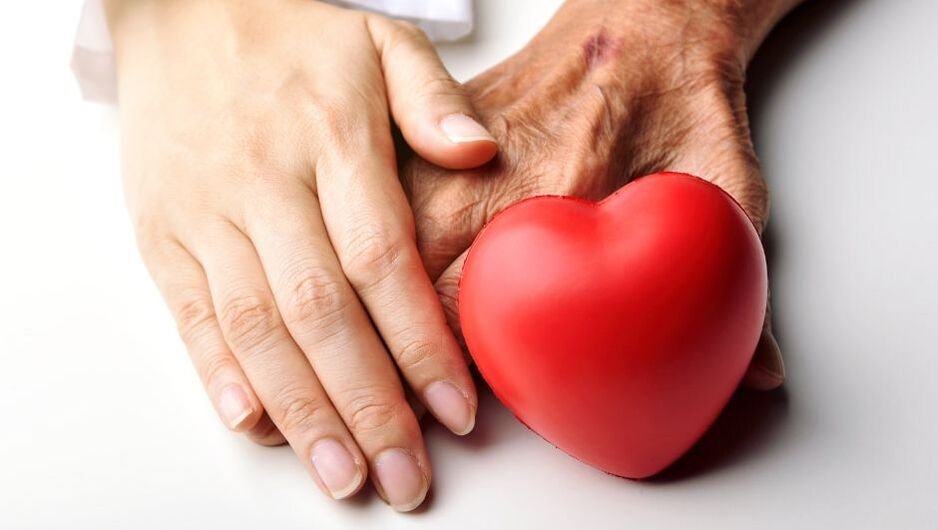 A young person's hand and an elderly person's hand holding each other with a red heart-shaped object nearby. Tuktu