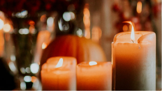 Lit orange and cream-colored candles on a table with blurred background of holiday lights and decorations. Anora Funeral Services. 