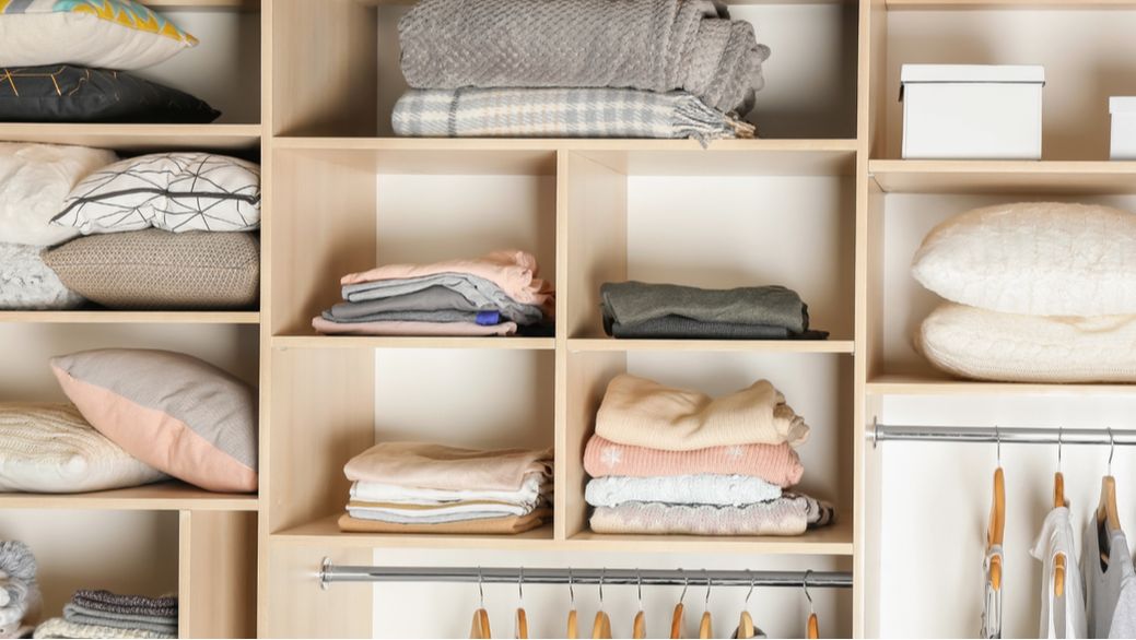 Open wooden closet with neatly folded clothes, pillows, and storage boxes. Good Riddance Organizers. 