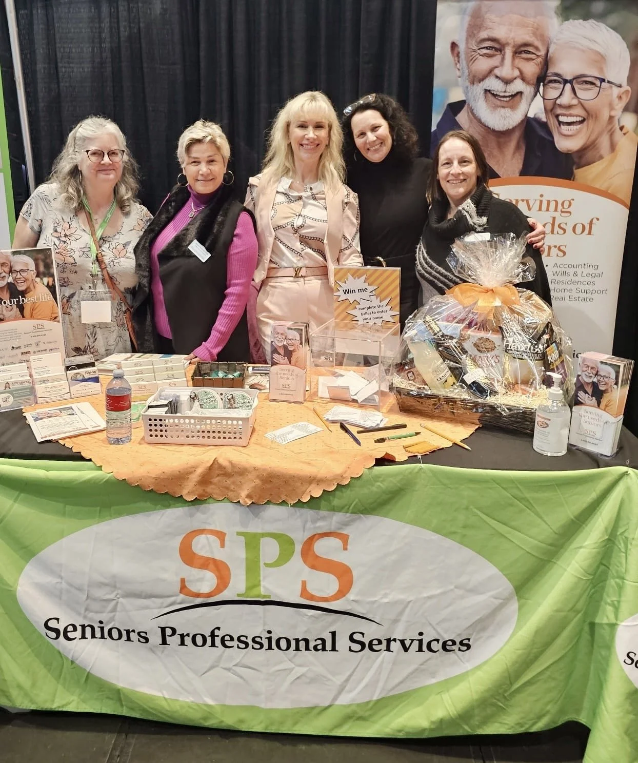 Five businesswomen standing behind a booth for Seniors Professional Services at an event. The table has informational materials. A vertical banner with smiling elderly people and the company's mission statement is visible in the background.