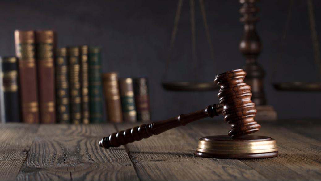 Gavel resting on sound block on a wooden table, with legal books and a balance scale in the background. HS Law Corporation. 