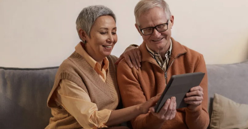 An elderly man and woman sitting on a sofa, looking at a tablet together and smiling.