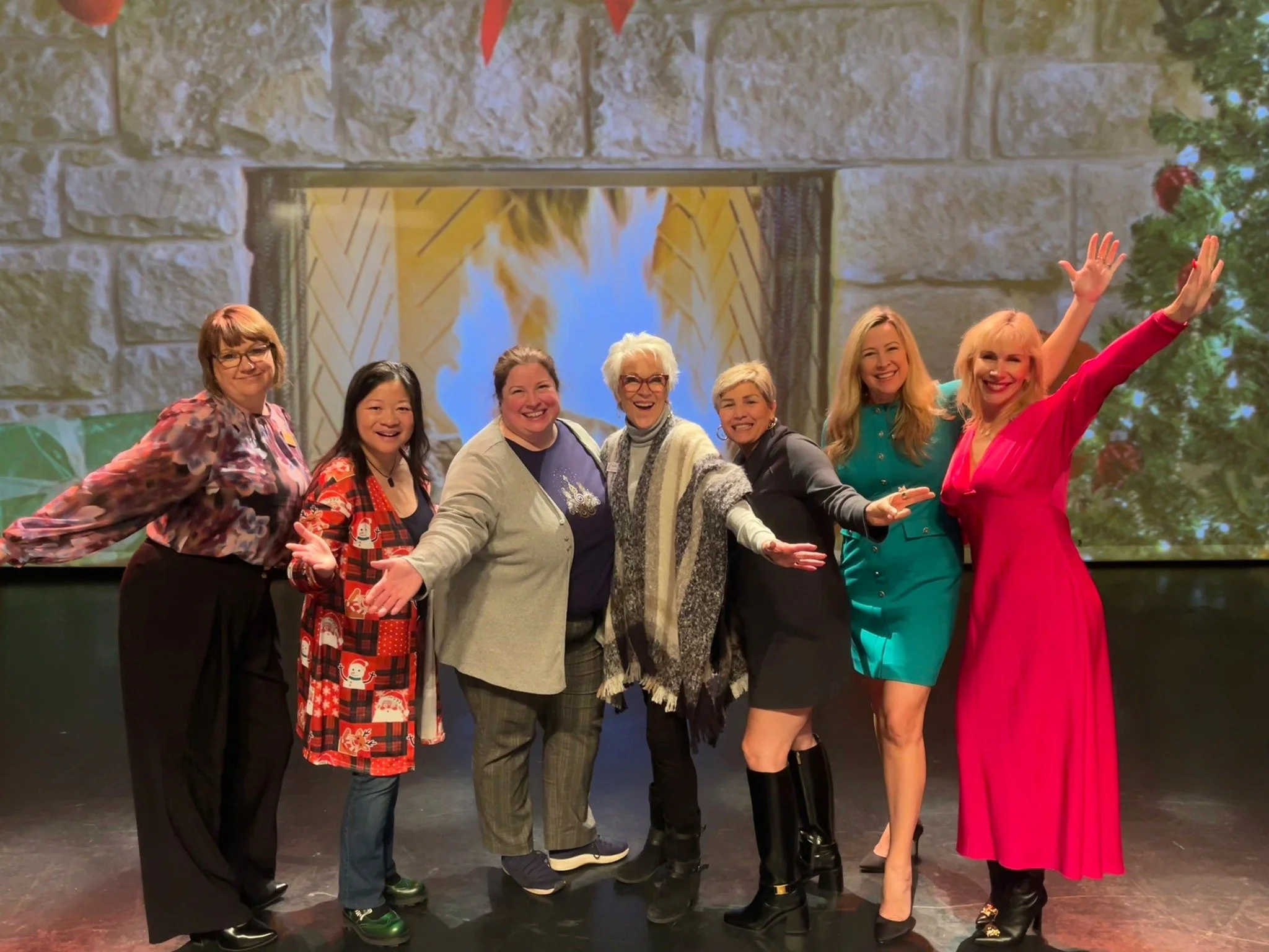 Seven women standing on stage, smiling and posing for a photo, with a backdrop featuring a fireplace and Christmas tree, at a Tri-Cities Chamber of Commerce event.
