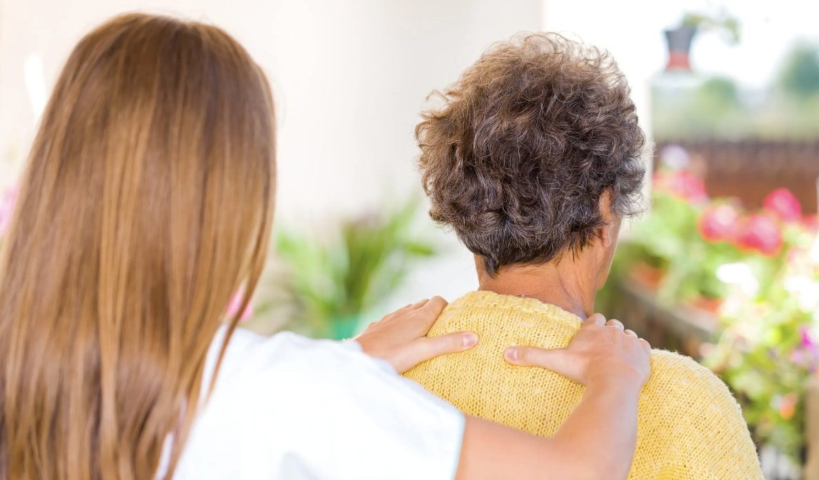 A healthcare professional providing support to an elderly woman with gray, curly hair, who is wearing a yellow sweater, in an indoor setting with blurred plants and flowers in the background.