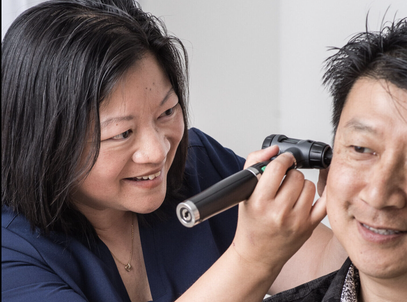 A woman with dark hair is examining a man's ear using an otoscope, in a clinical setting. Network Hearing in Port Coquitlam. 