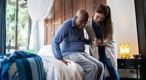 A man sitting on a bed looking at a smartphone with a woman standing next to him in a room with wooden walls and a window. Representing Assisted living, home care support. 
