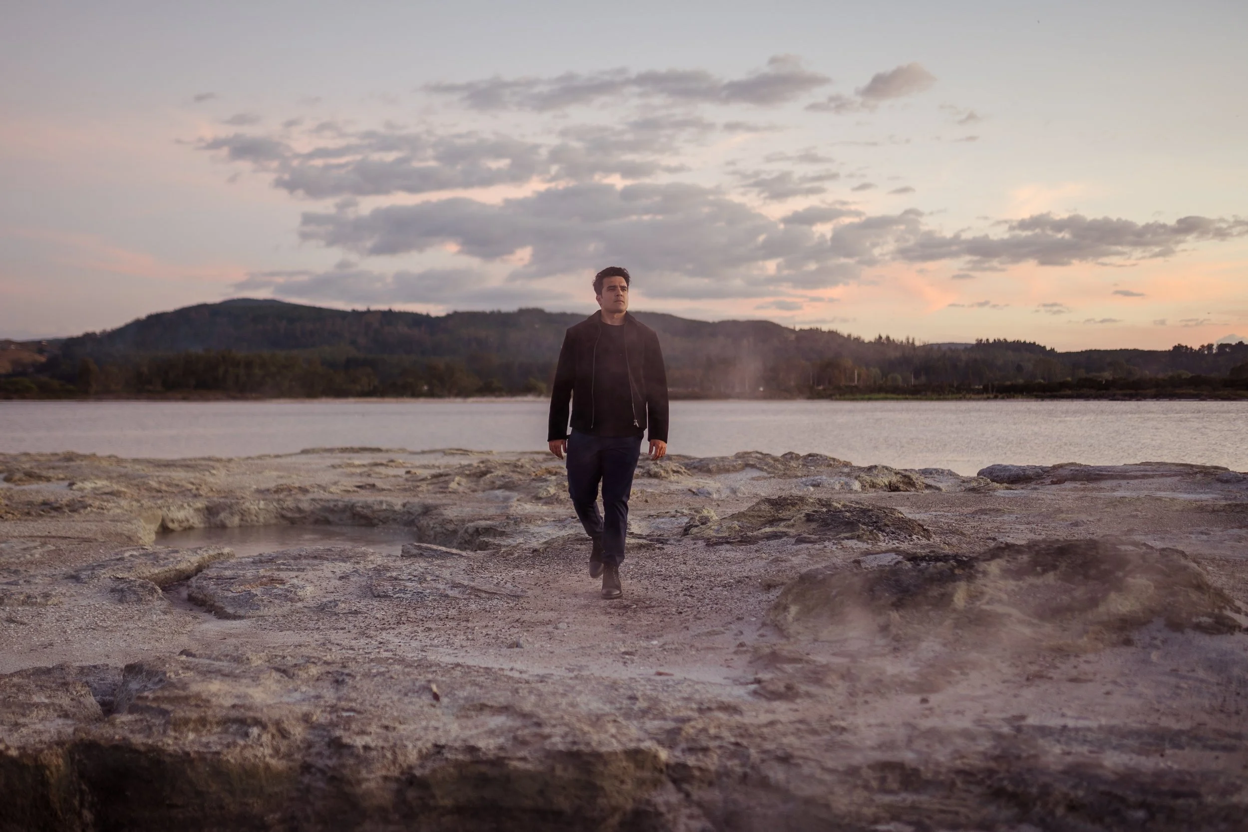 A man walking on rocky terrain near a body of water at sunset or sunrise, with hills and a cloudy sky in the background.