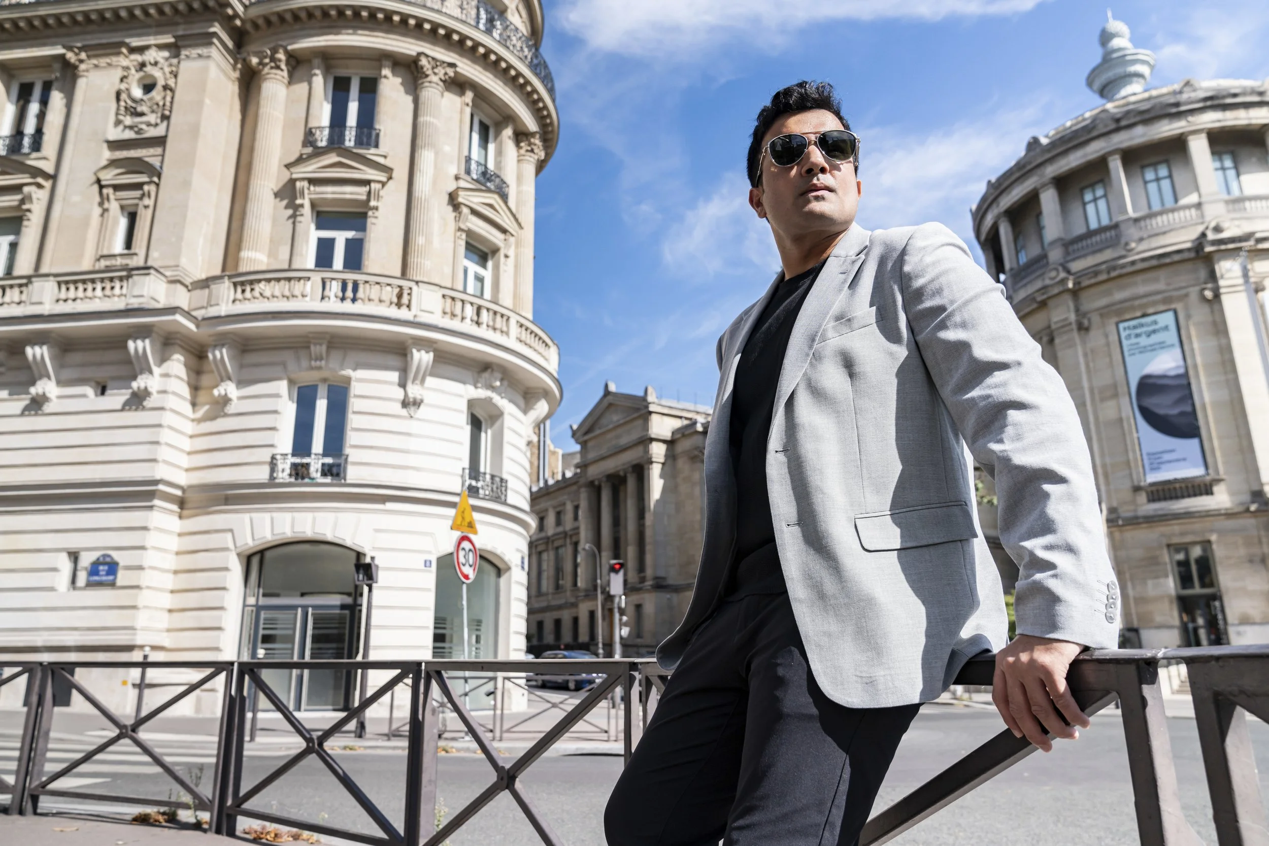 A man wearing sunglasses and a light gray blazer leaning against a black metal fence on a city street, with historic buildings and a blue sky in the background.