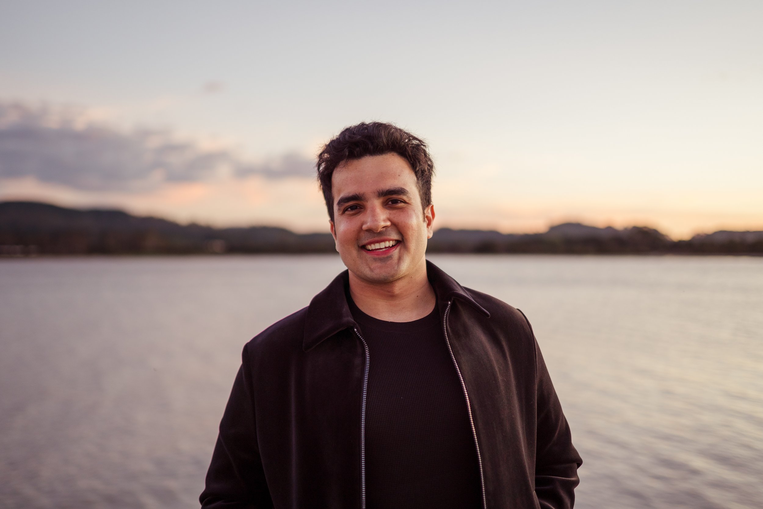 A young man with dark, curly hair and light skin smiling outdoors near a body of water during sunset, wearing a black jacket and black shirt.