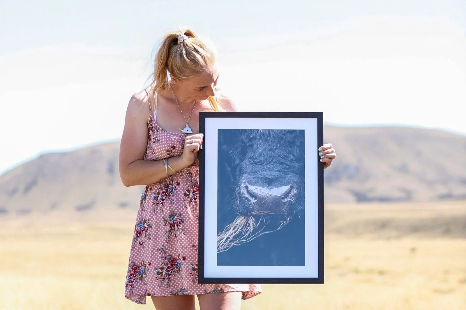 A woman in a pink floral dress standing outdoors in a field, holding a framed photograph of a lion's face.