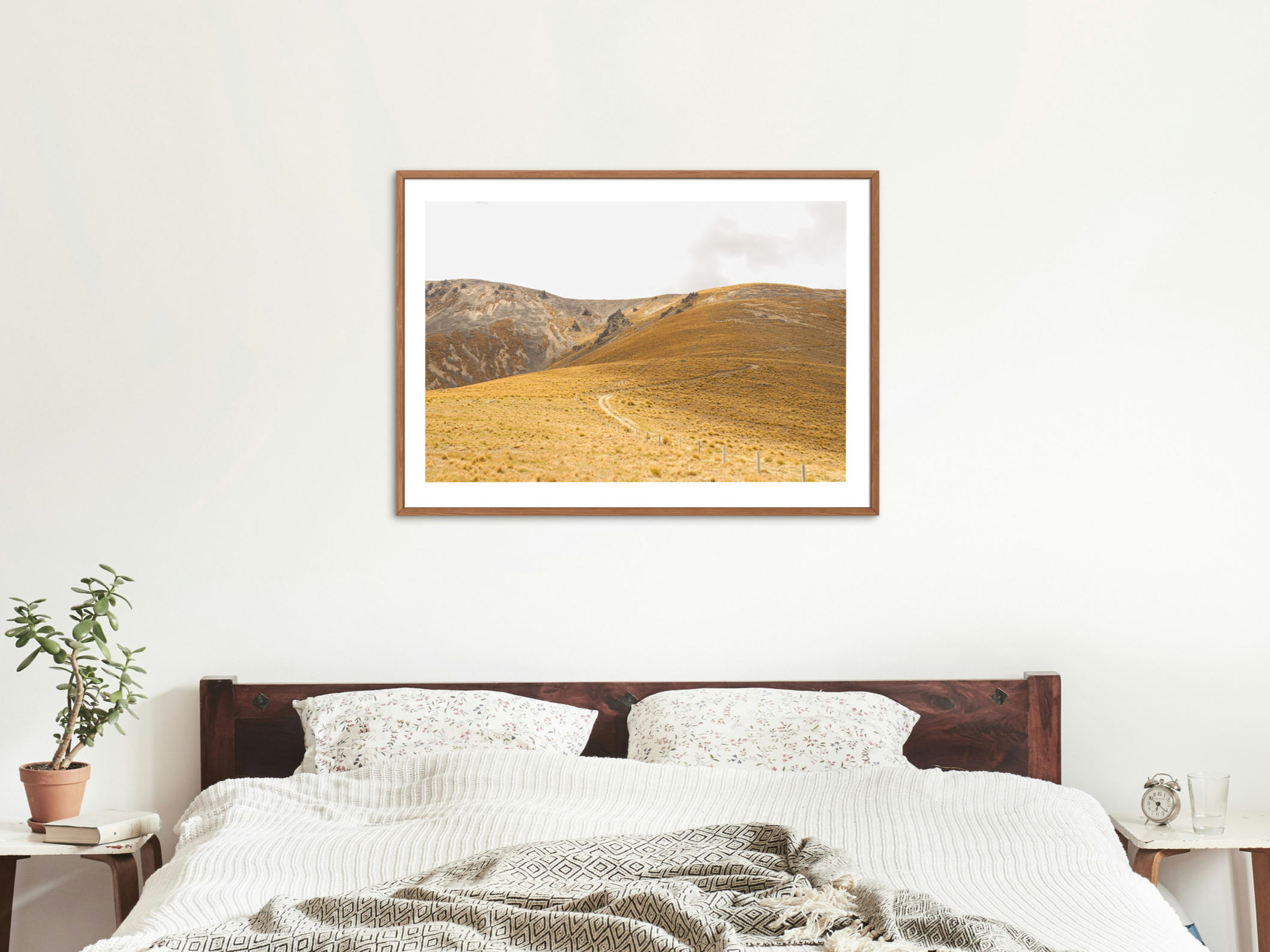 A framed landscape photograph of a mountain scene hangs above a bed with white and patterned pillows and a beige blanket in a bedroom with white walls, a potted plant, and bedside tables.