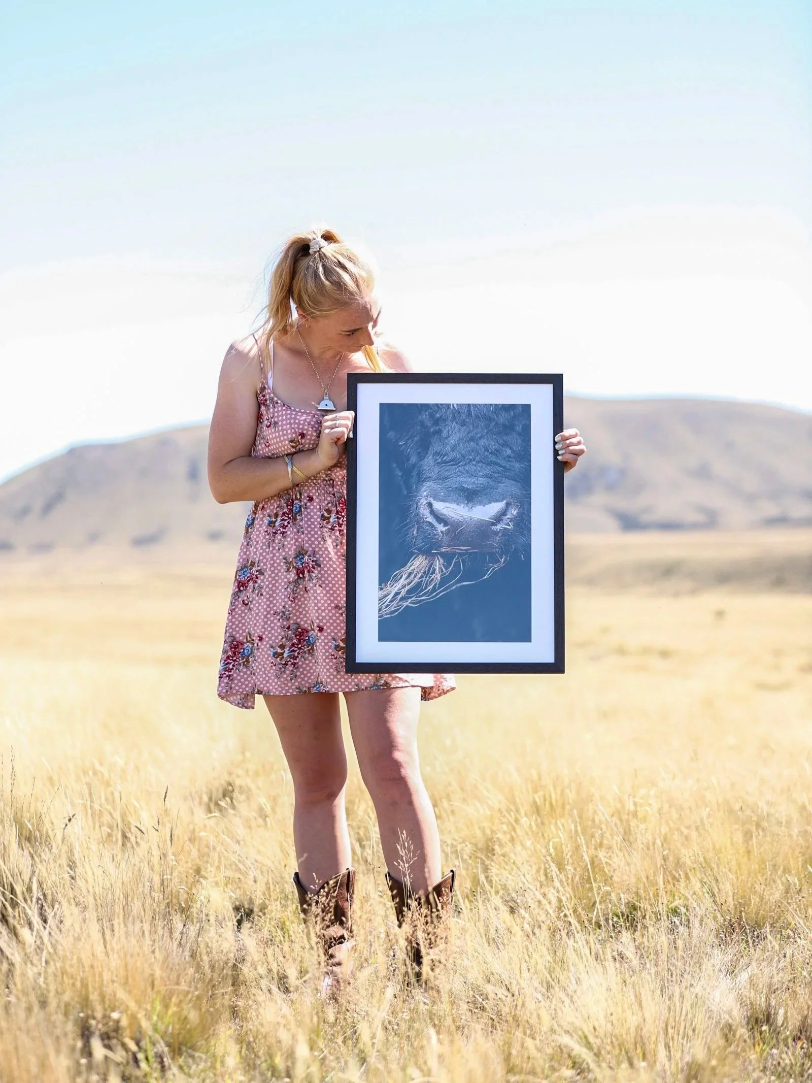 A woman standing in a grassy field holding a framed photograph of a lion's face, with mountains in the background.