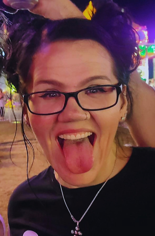 A young woman with glasses sticking out her tongue at night in a lively outdoor setting with colorful lights.