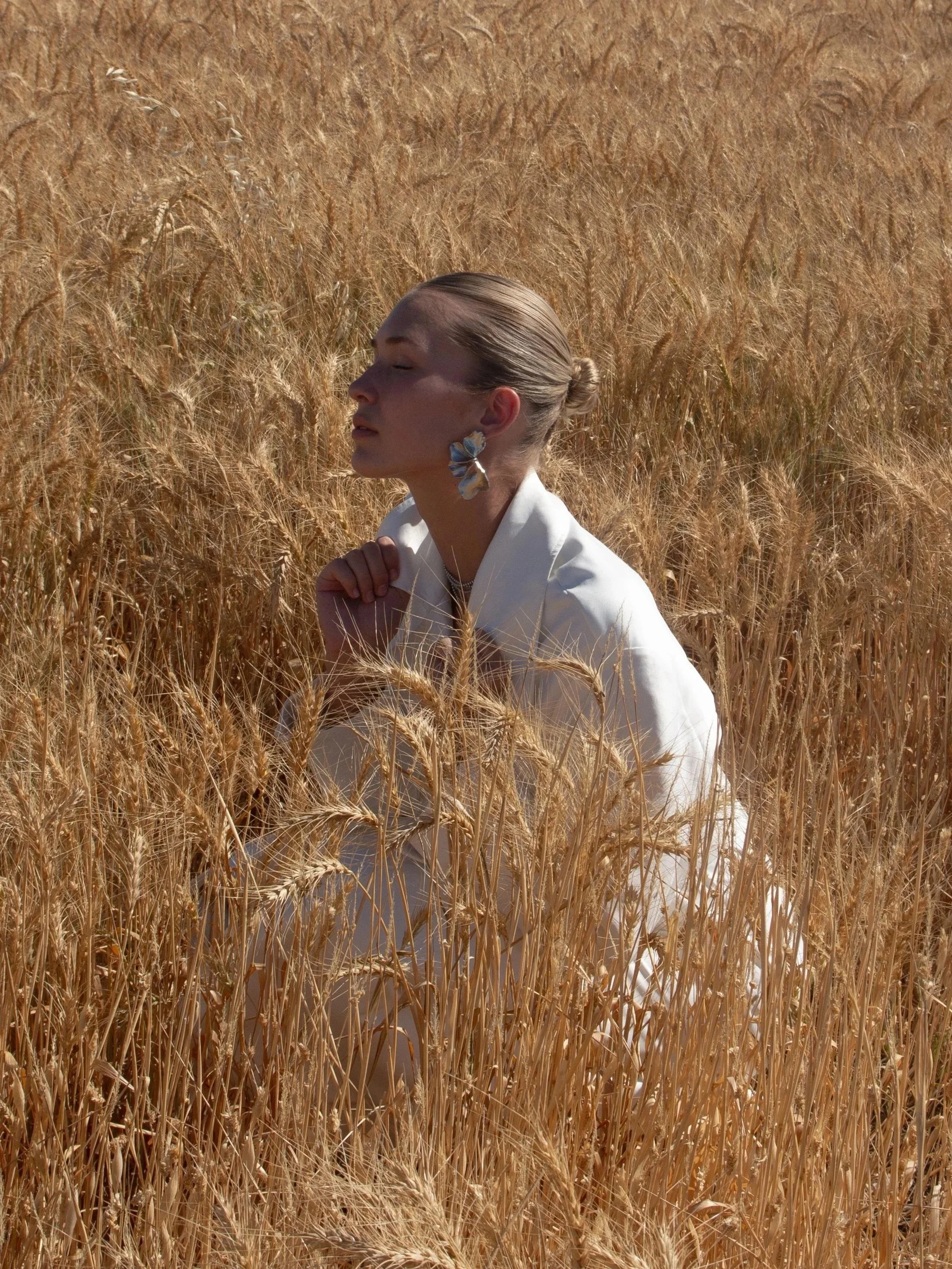 A woman with closed eyes and a bun hairstyle, wearing white clothing and large butterfly-shaped earrings, sitting in a golden wheat field.