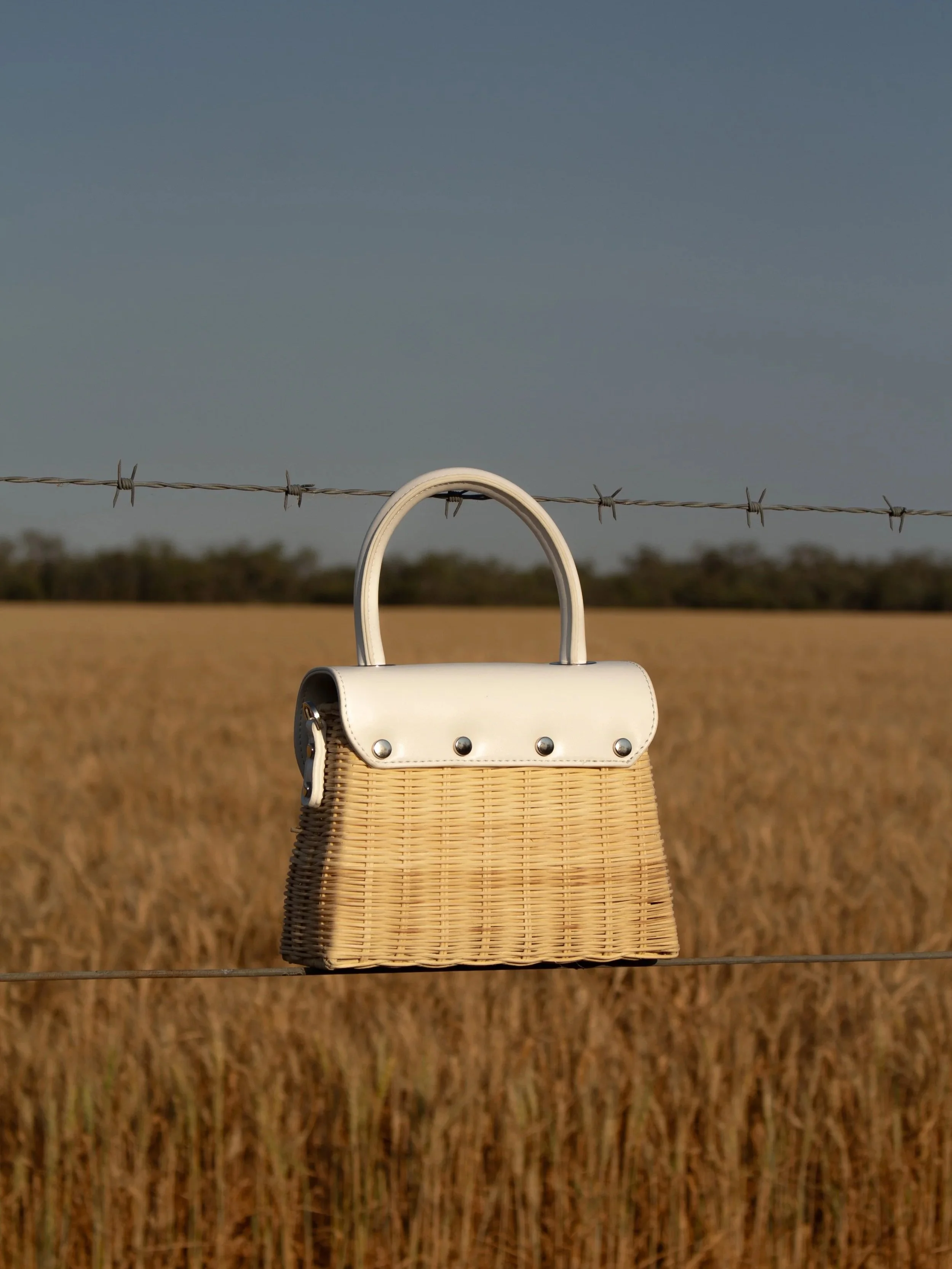 A woven handbag with a white leather top and handle hanging on a barbed wire fence in a field.