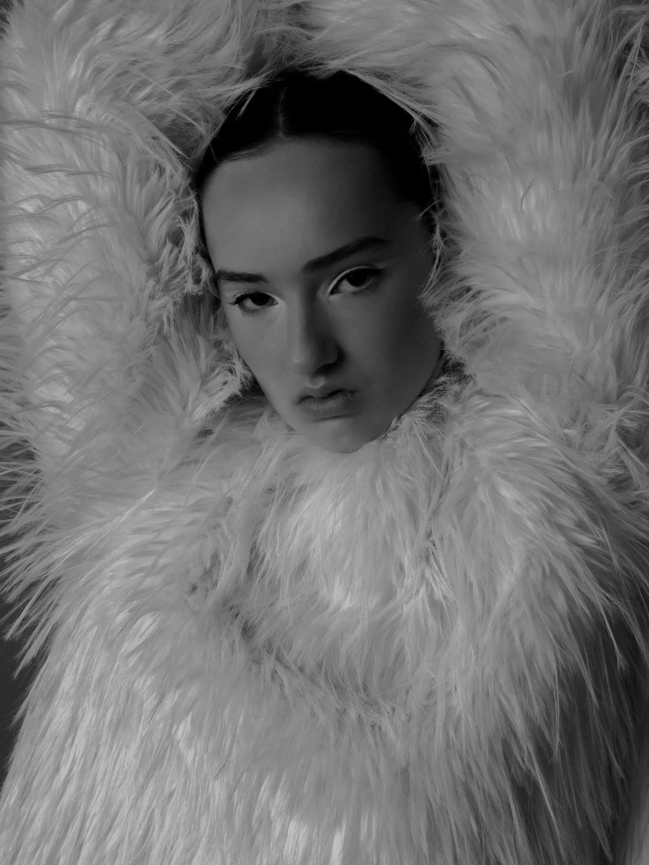 Black and white portrait of a woman with dark hair, surrounded by fluffy fur, gazing directly at the camera.