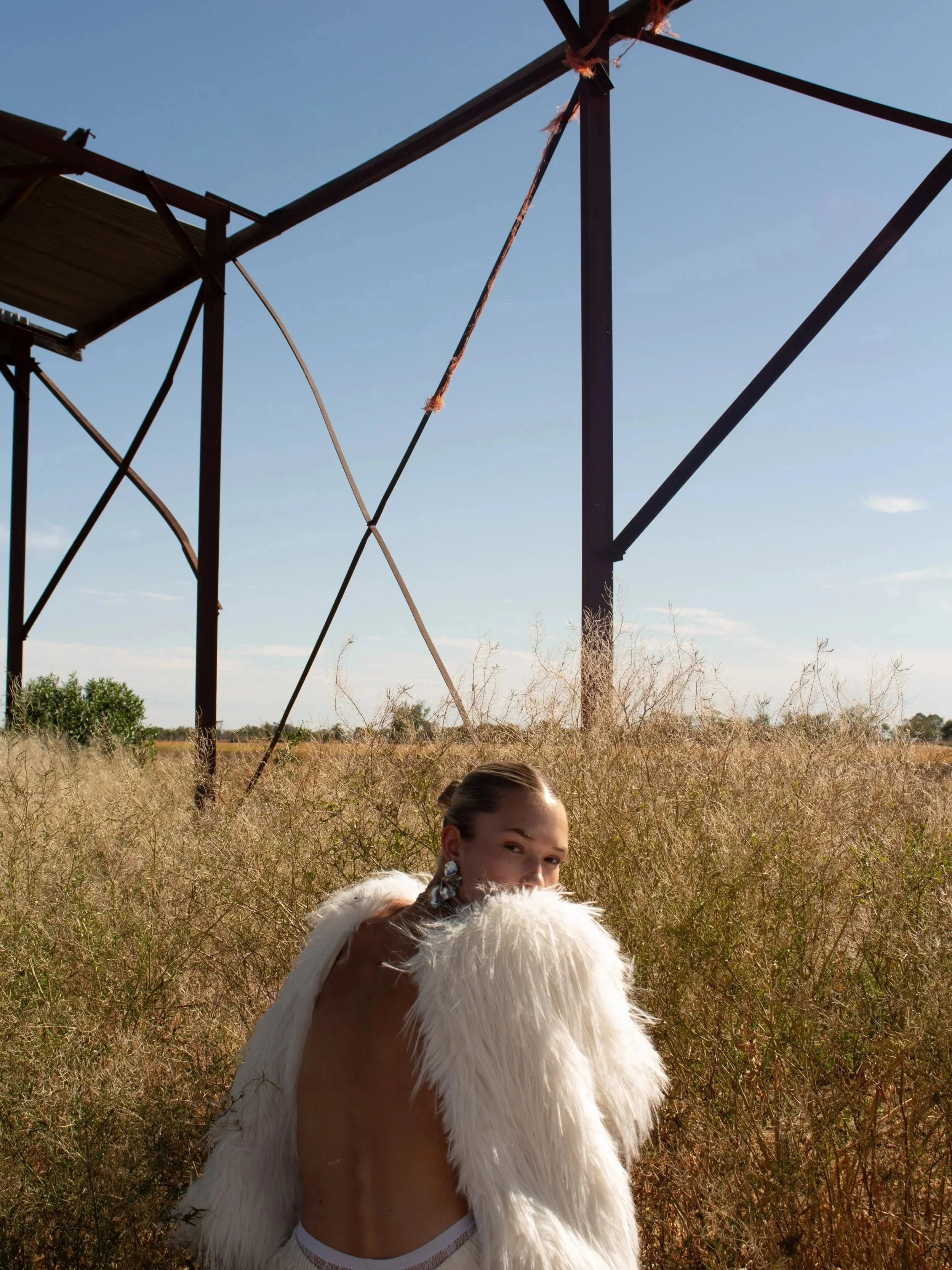 A woman with a high bun hairstyle and earrings wearing a white fluffy fur coat, standing in a dry grassy field under a clear blue sky, with a large metal tower structure behind her.