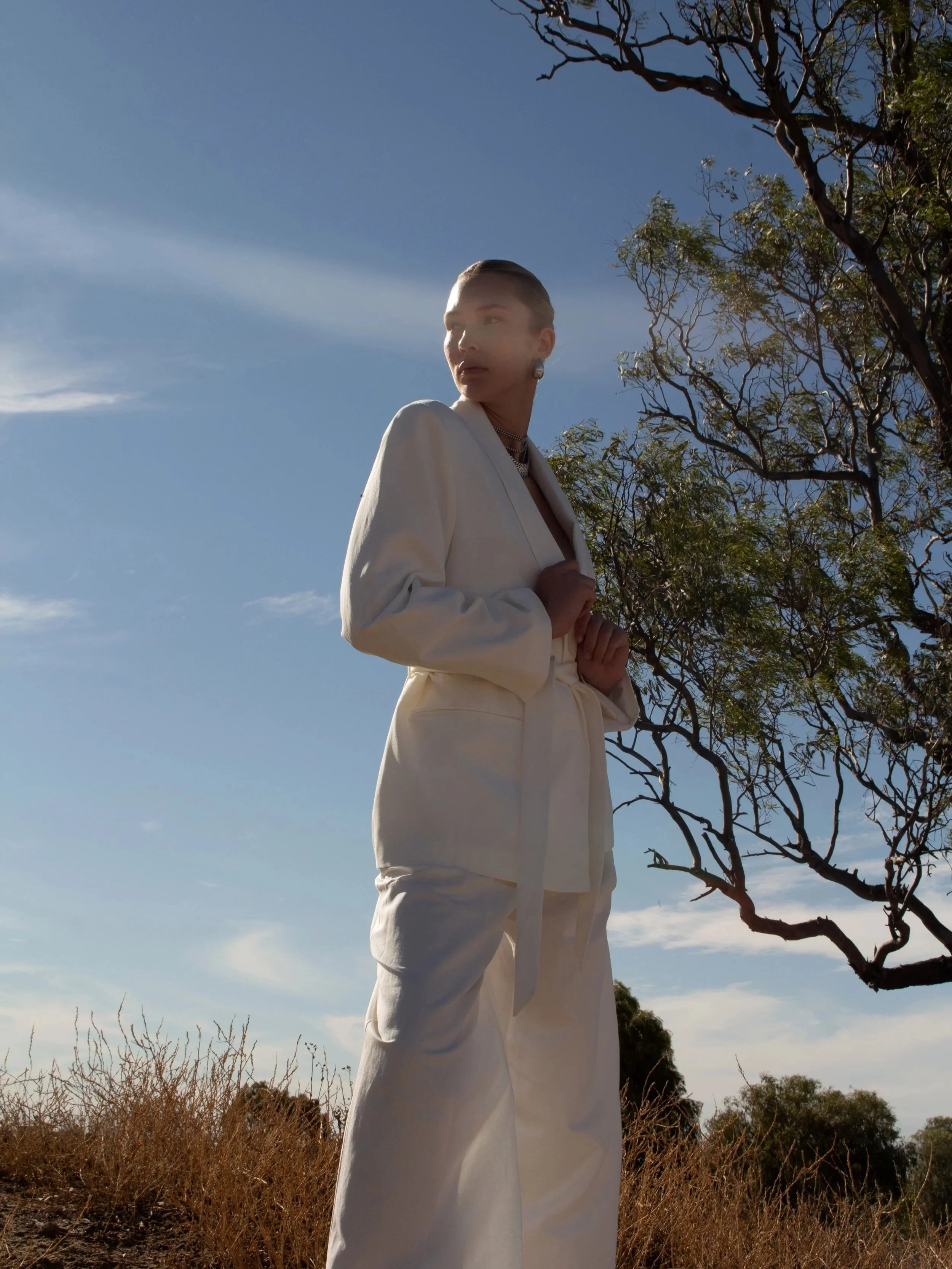 A woman standing outdoors during daytime, dressed in a white suit with a belt, with her hair slicked back, jewelry, and jewelry, against a backdrop of trees and a blue sky with some clouds.