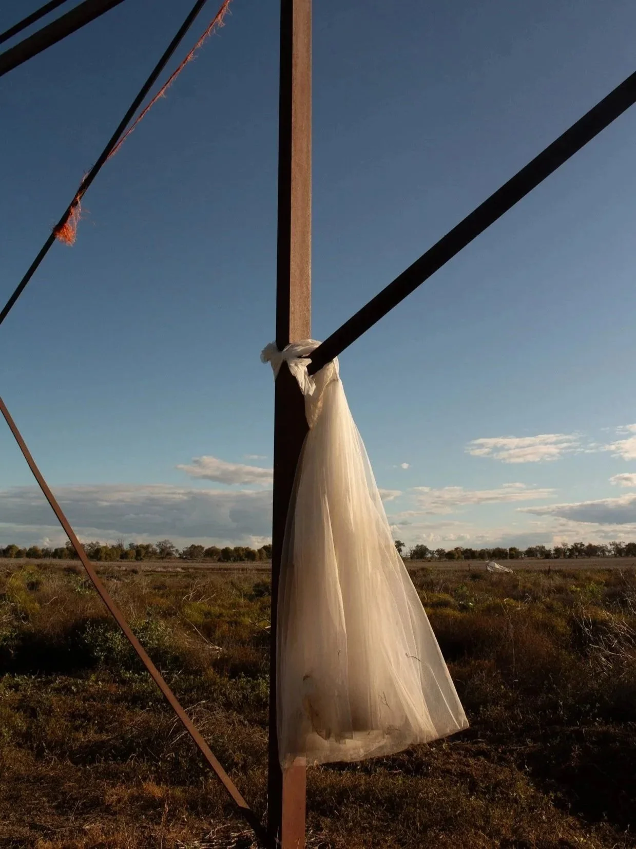 A wedding veil tied to a wooden post with black ropes, set in a rural landscape under a blue sky with scattered clouds.