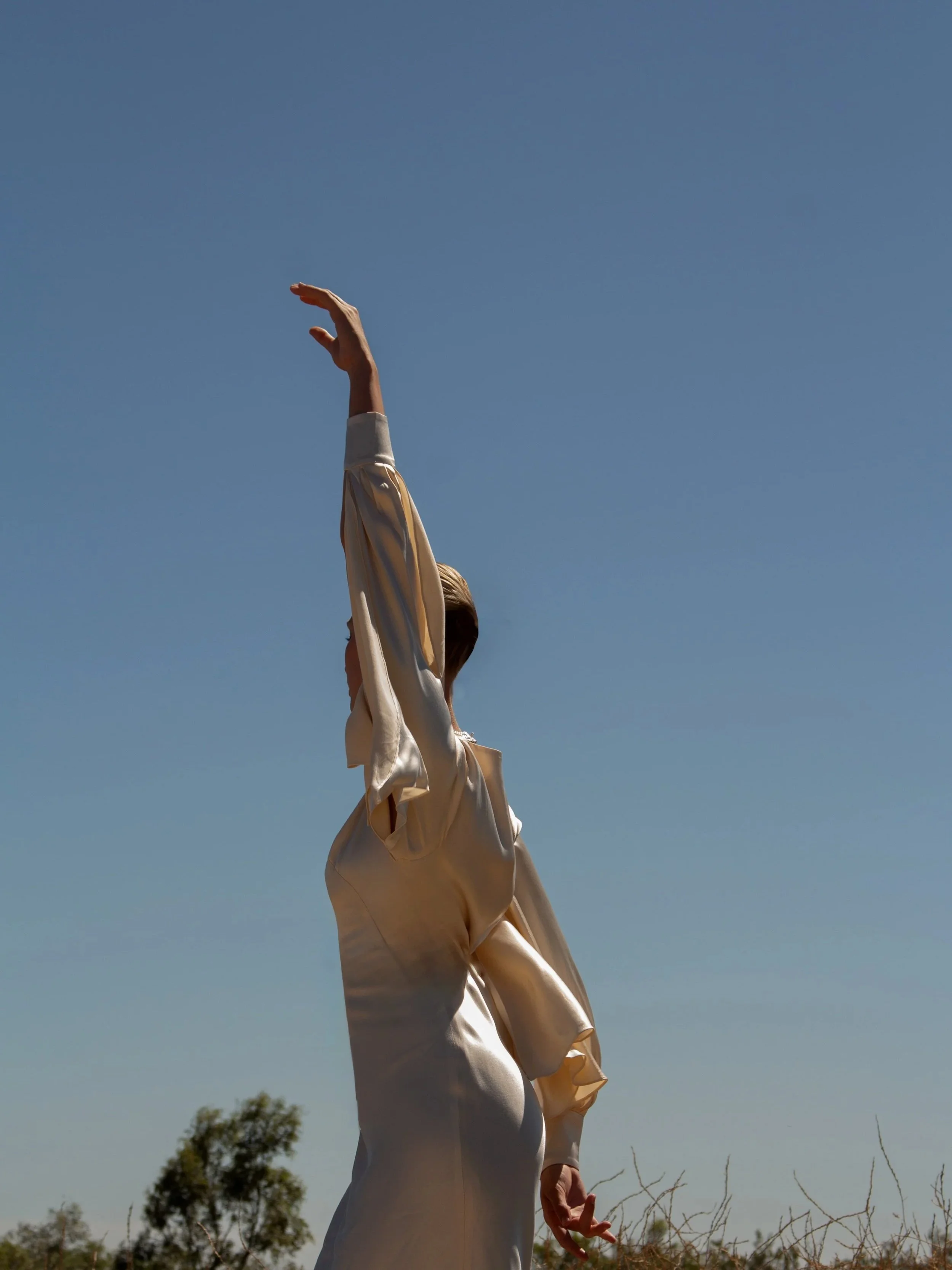 A person is standing outdoors against a clear blue sky, wearing a shiny, light-colored outfit, with one arm raised above their head and the other hanging down.