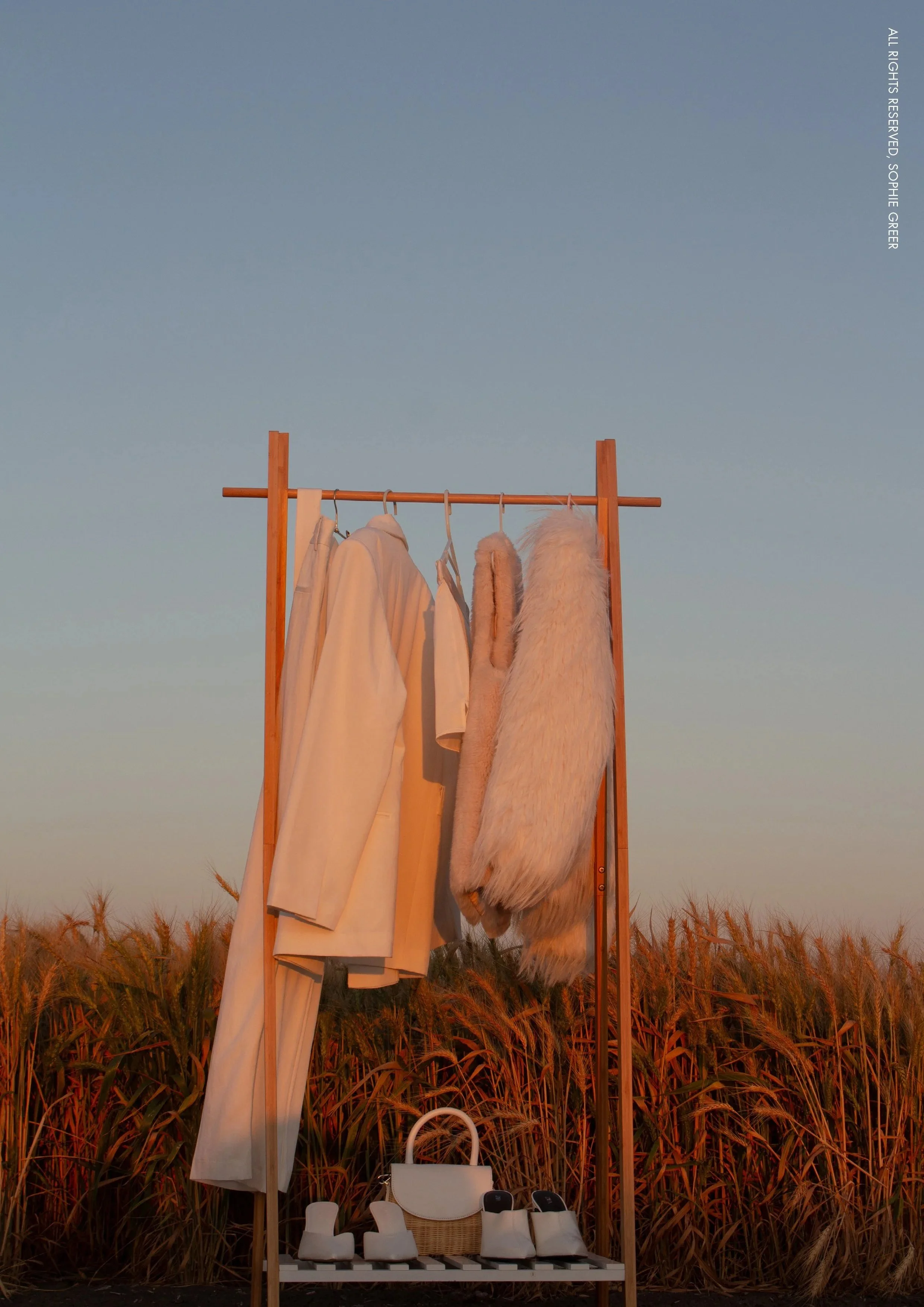 Clothing rack with white coats, furry beige coat, and handbags, set against a wheat field during sunset.