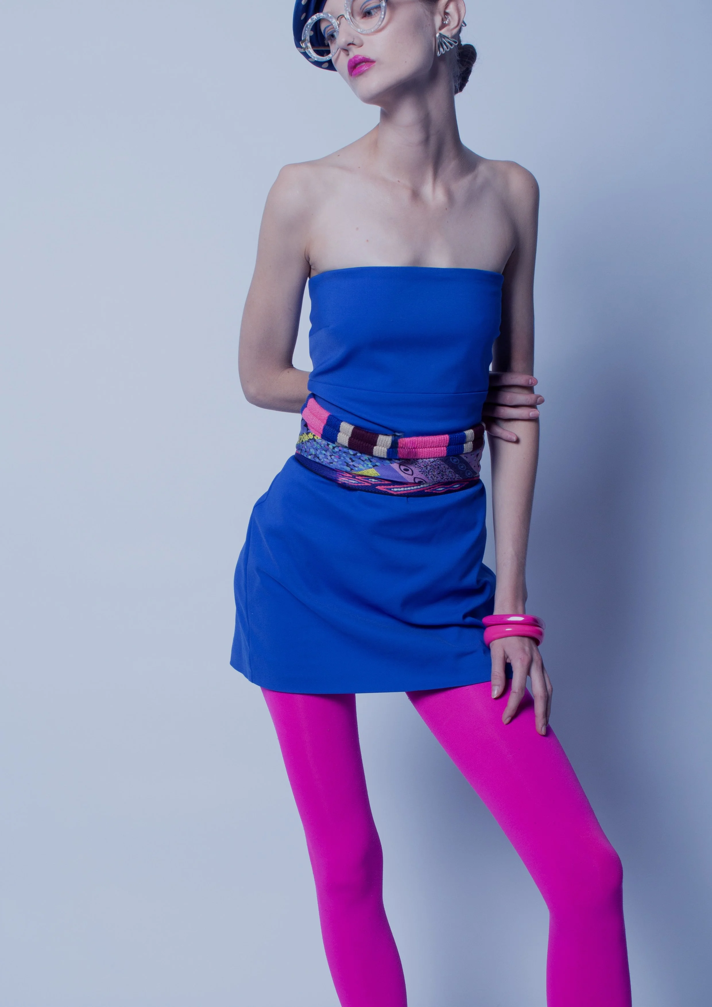 A woman with short hair and glasses wearing a blue strapless dress, pink tights, and pink accessories, posed against a plain wall.