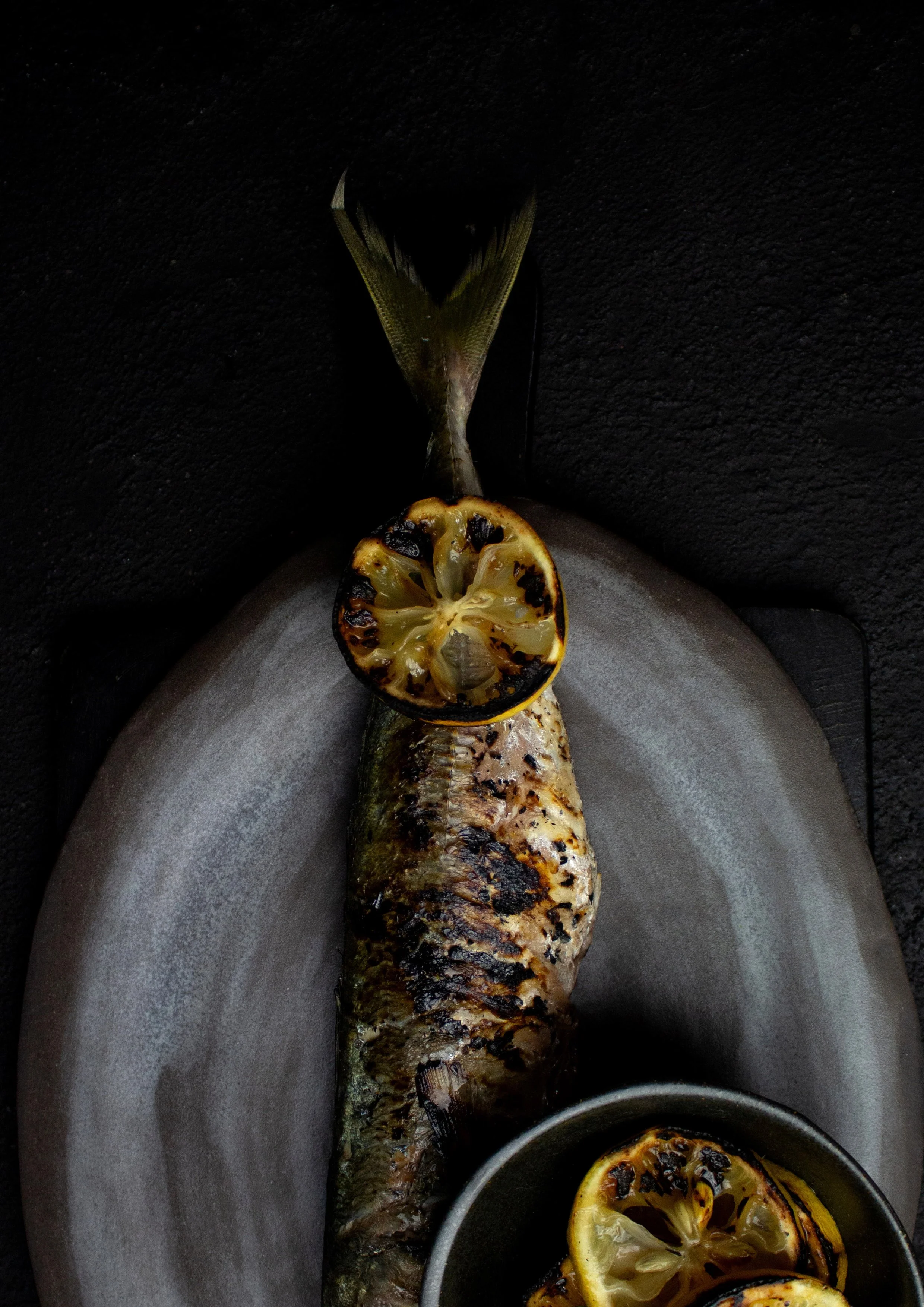 Grilled fish with lemon slices on a dark plate.