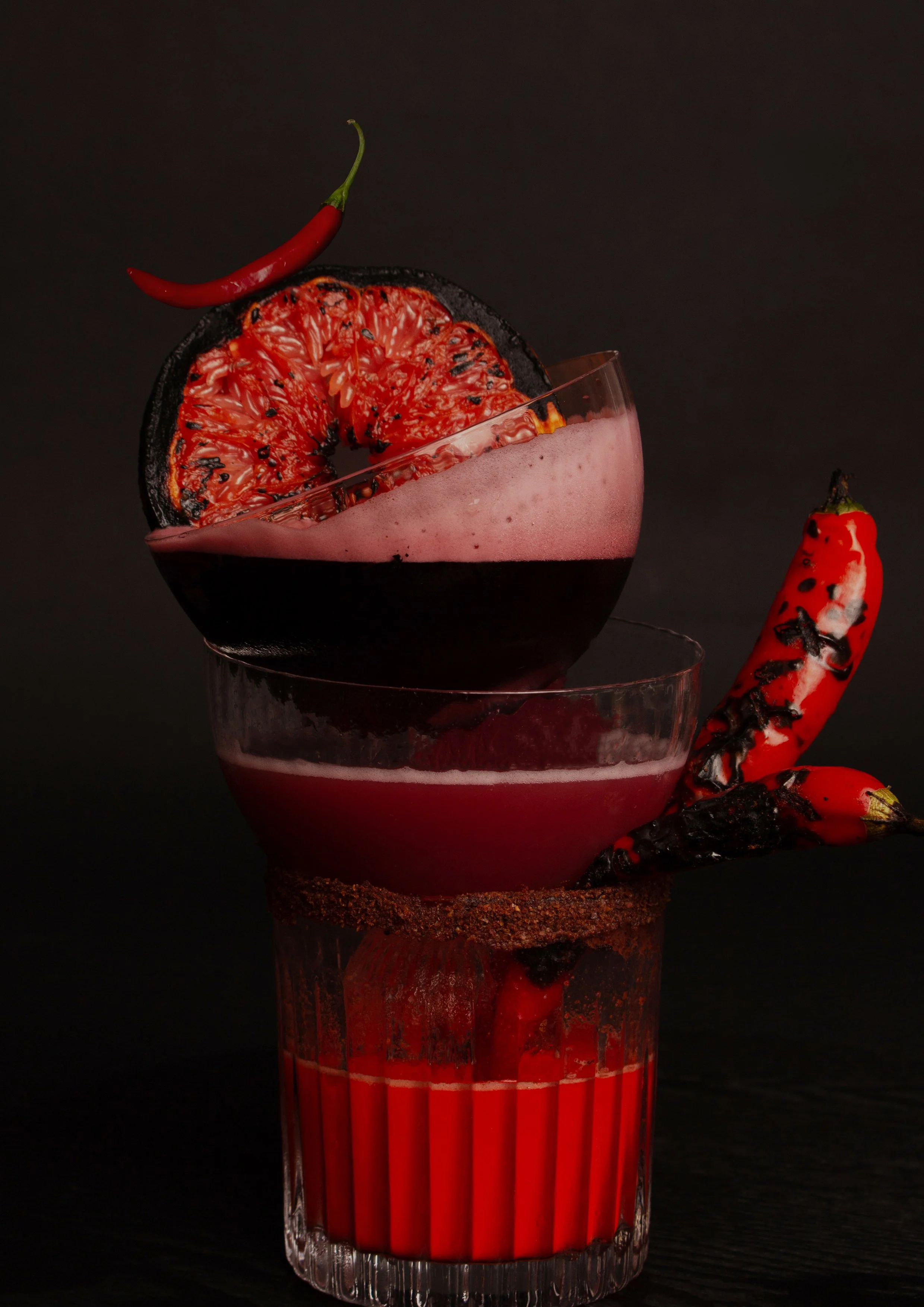 A multi-layered cocktail with a dark red base in a textured glass, topped with a pink foam layer in a martini glass, garnished with charred citrus slices and red chili peppers.