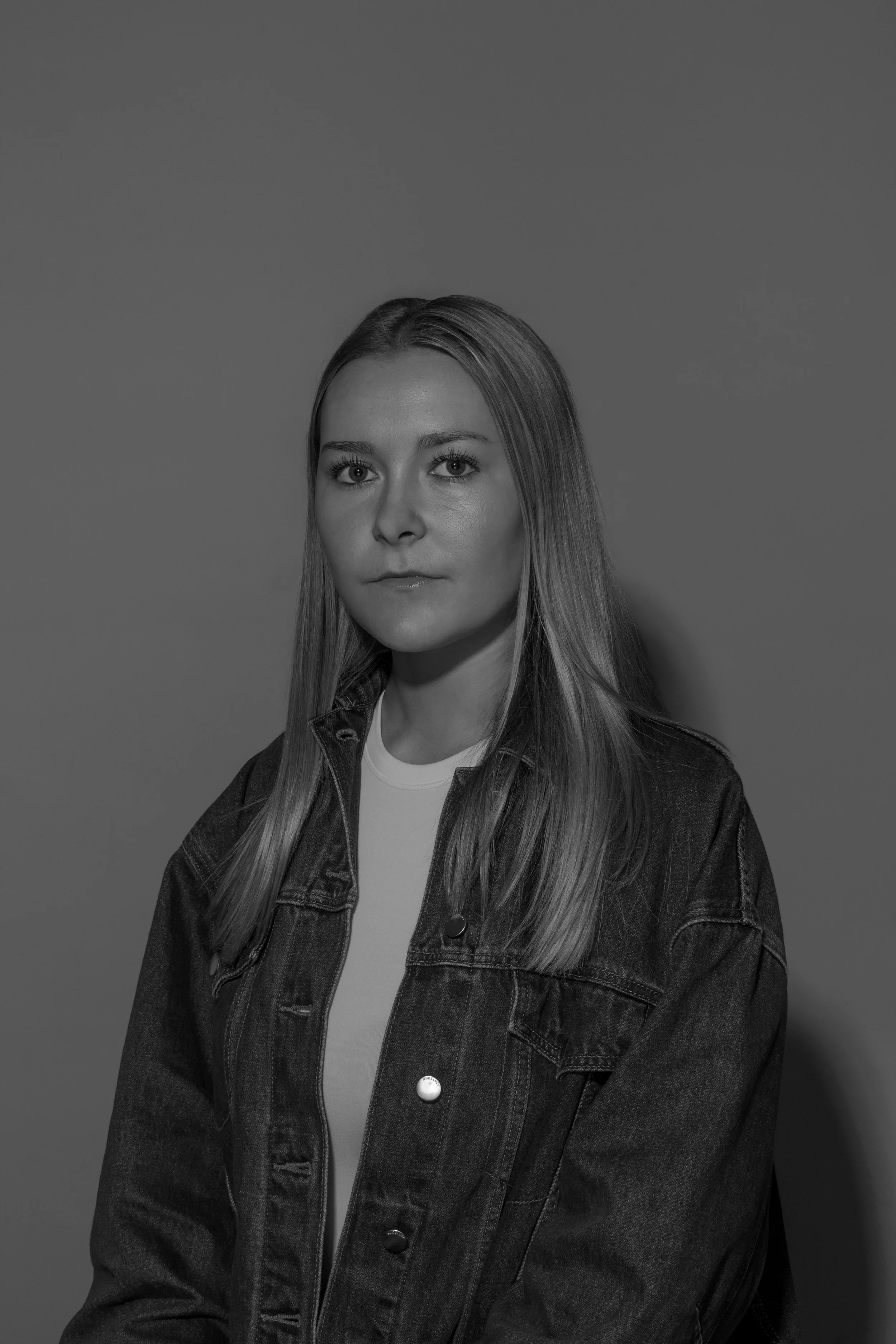 Black and white portrait of a young woman with straight hair, wearing a denim jacket over a light shirt, against a plain background.