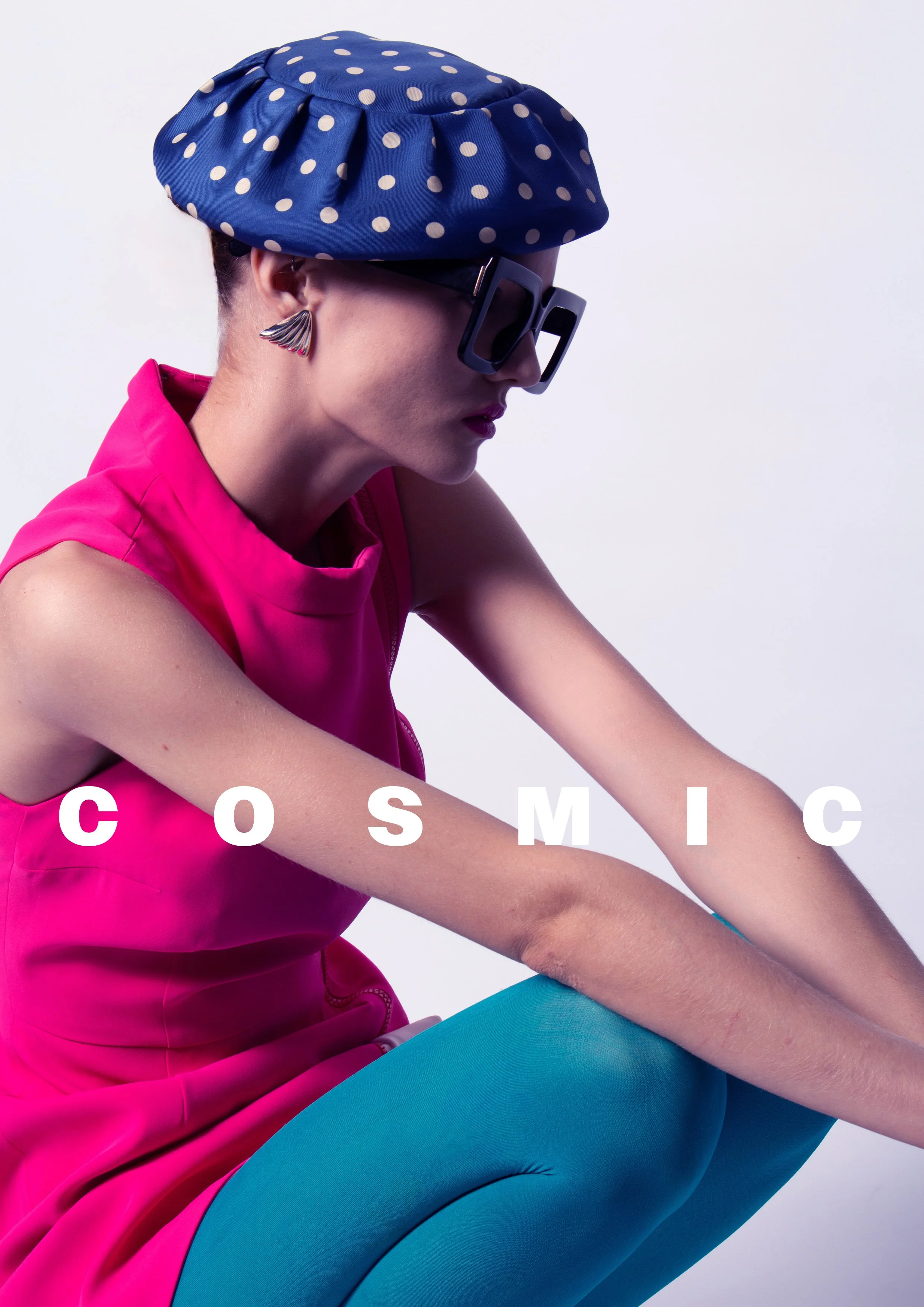 A woman dressed in bright pink, blue tights, wearing a large blue polka-dot hat, oversized black glasses, and colorful earrings, sitting against a plain light background with the word 'COSMIC' overlaid.