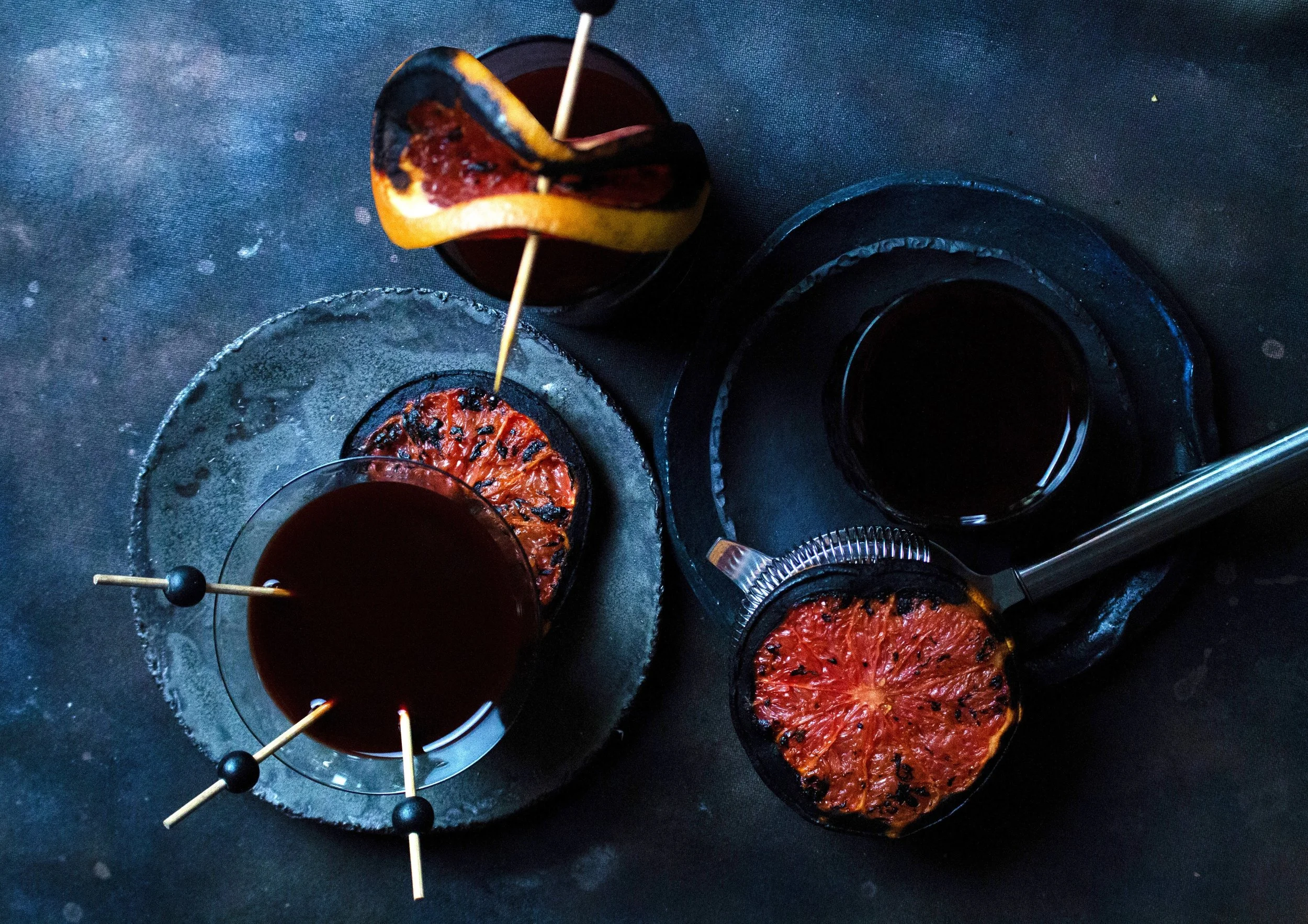 Top-down view of a black slate serving tray with grilled blood oranges and two glasses of dark red beverage, garnished with grilled orange slices on skewers.