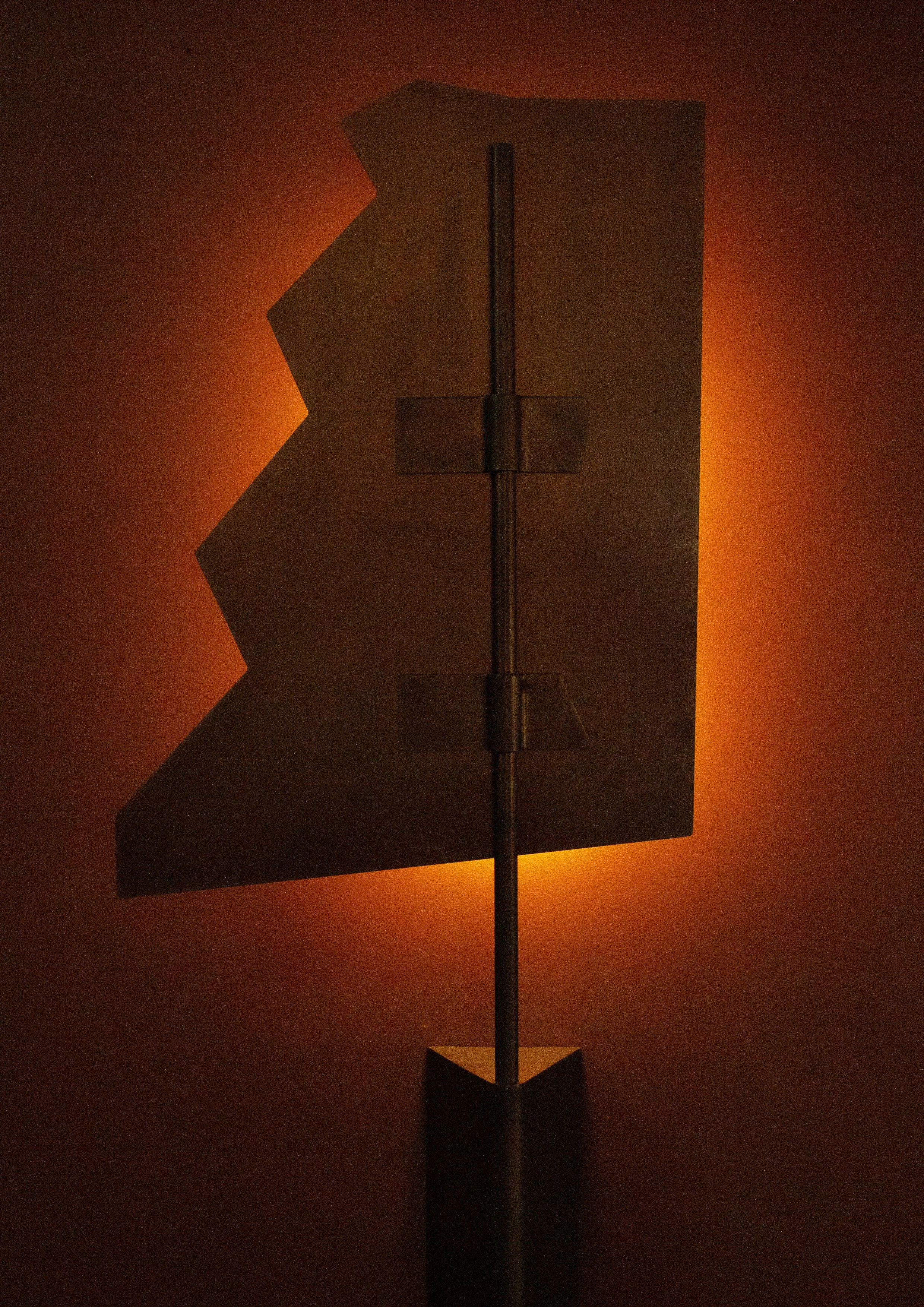 Backlit photograph of a metal flag or banner with a partially zigzag-edged shape, mounted on a stand against a dark background with warm orange glow.