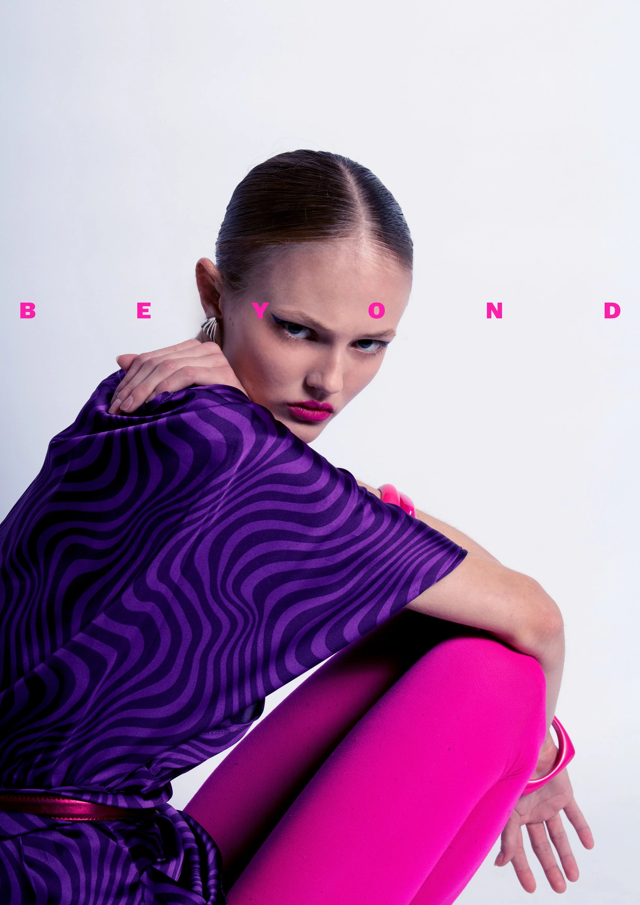 A woman with slicked-back hair wearing a purple striped top and bright pink tights, sitting with her knee up and hand resting on her knee, against a white background with pink letters spelling 'BEYOND' above her.