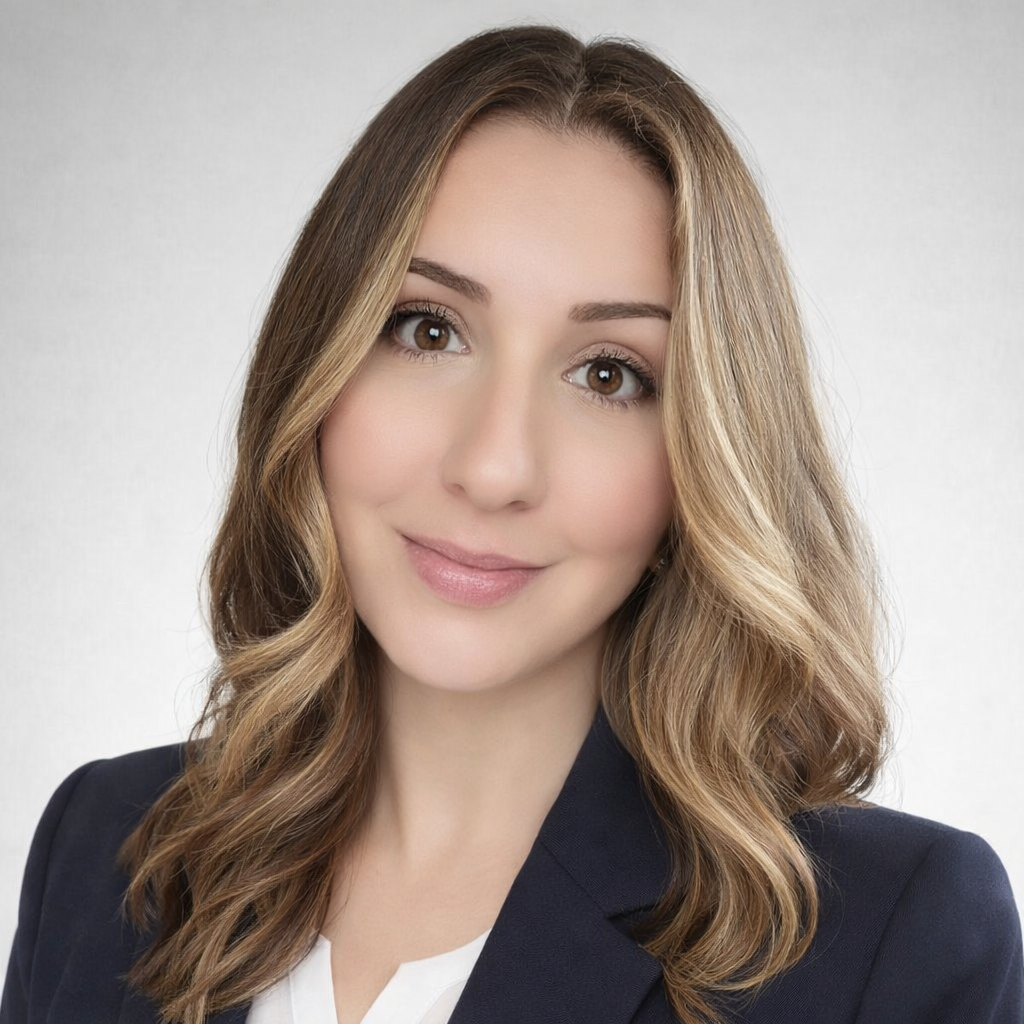 A woman with shoulder-length wavy hair, wearing a dark blazer and a white top, smiling softly against a light background.