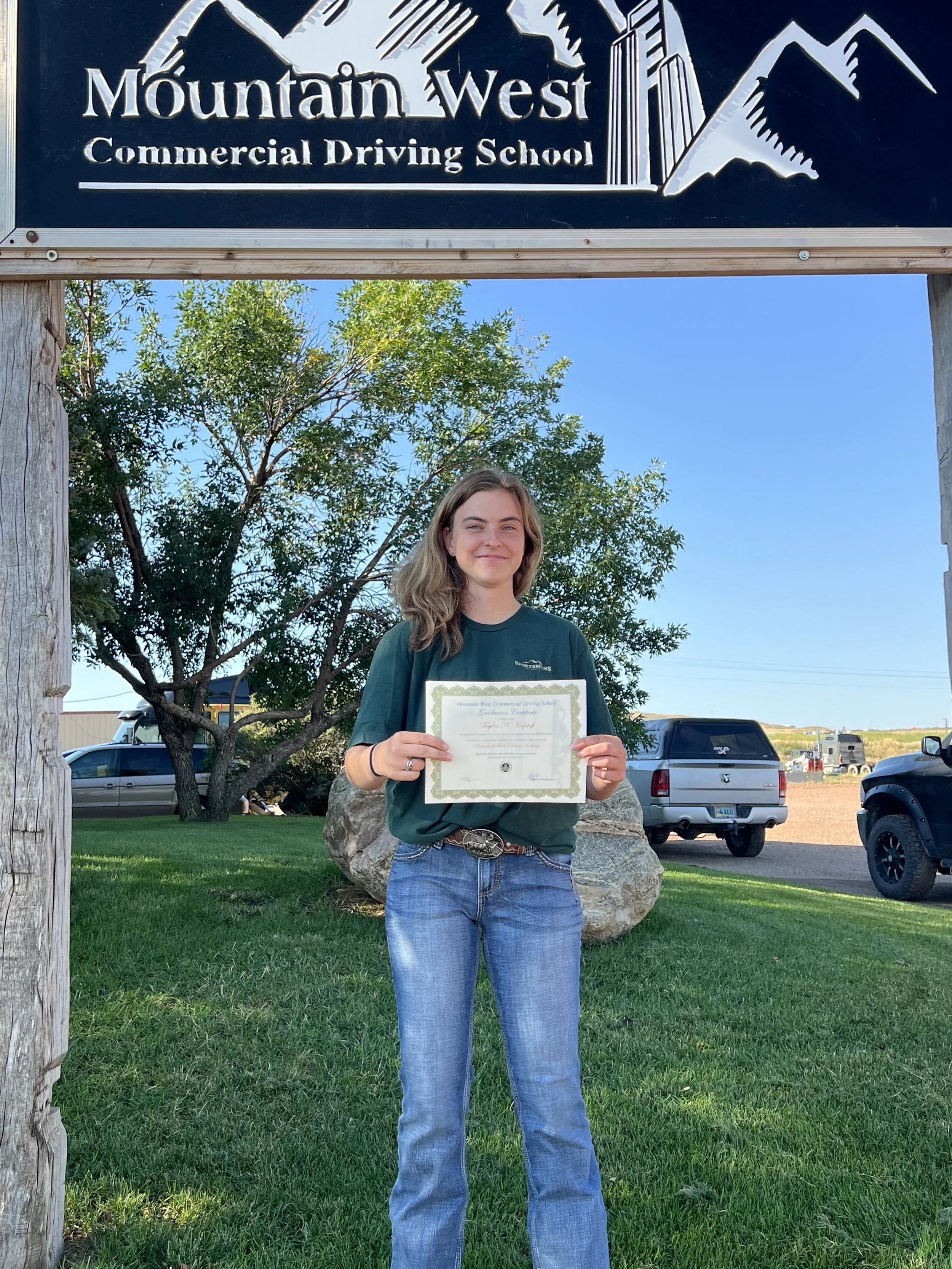 A young woman standing outside holding a certificate in front of a sign that reads 'Mountain West Commercial Driving School.' She is smiling and wearing a green t-shirt and blue jeans, with trees and parked cars in the background.