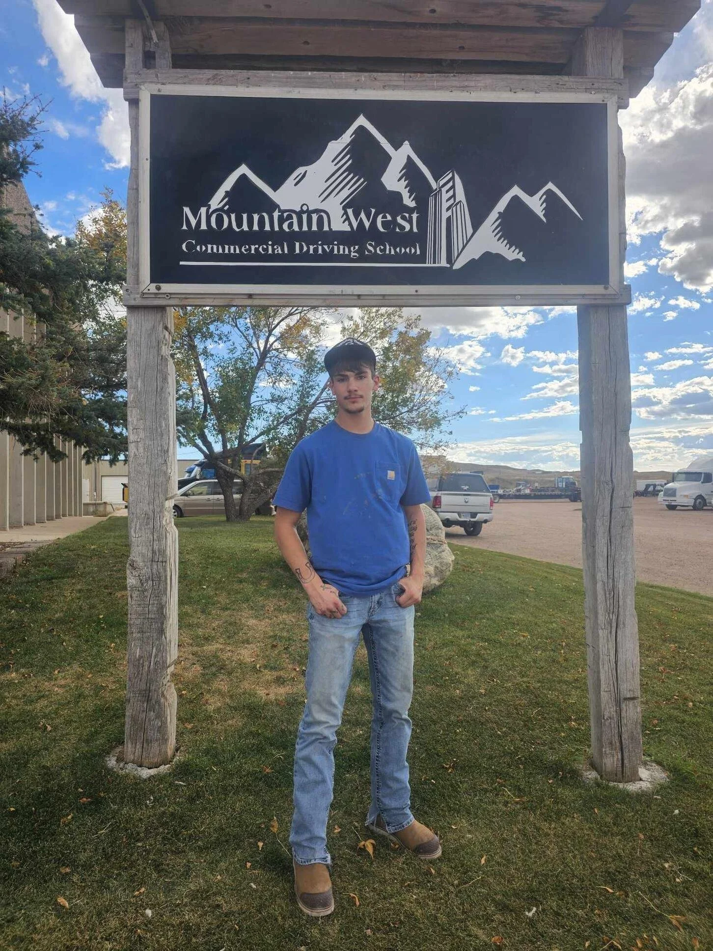 A young man wearing a blue t-shirt, jeans, and brown boots standing in front of a sign for Mountain West Commercial Driving School under a partly cloudy sky.