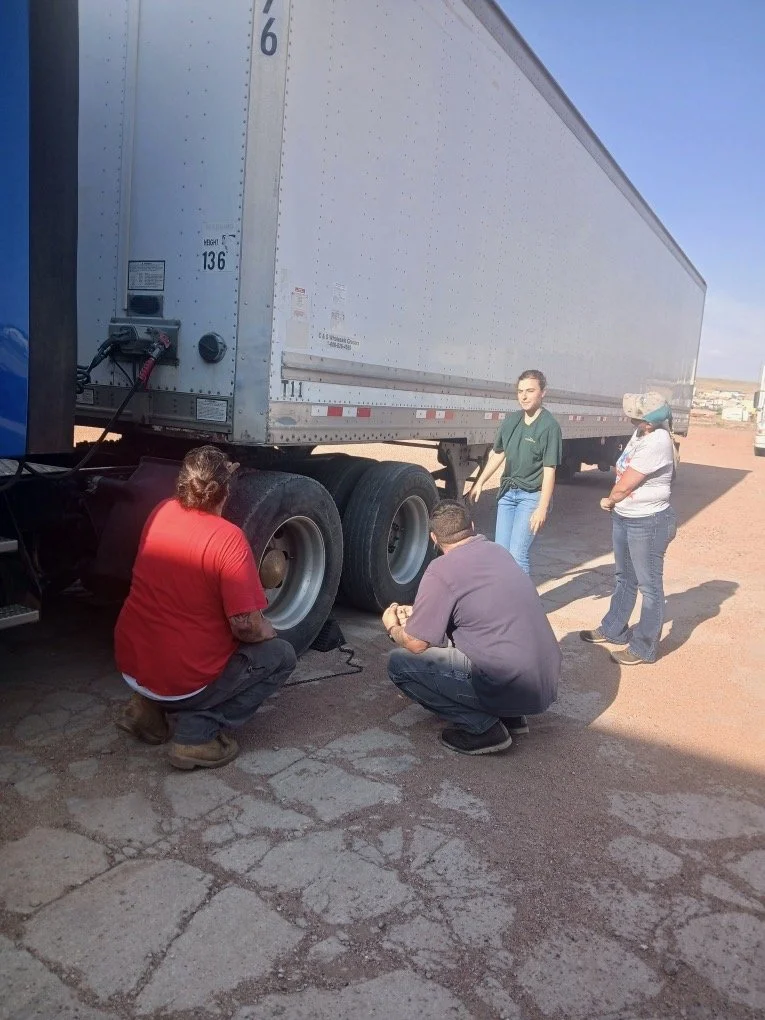 Four people working on the wheels of a large semi-truck trailer parked on a cracked pavement under a clear blue sky.
