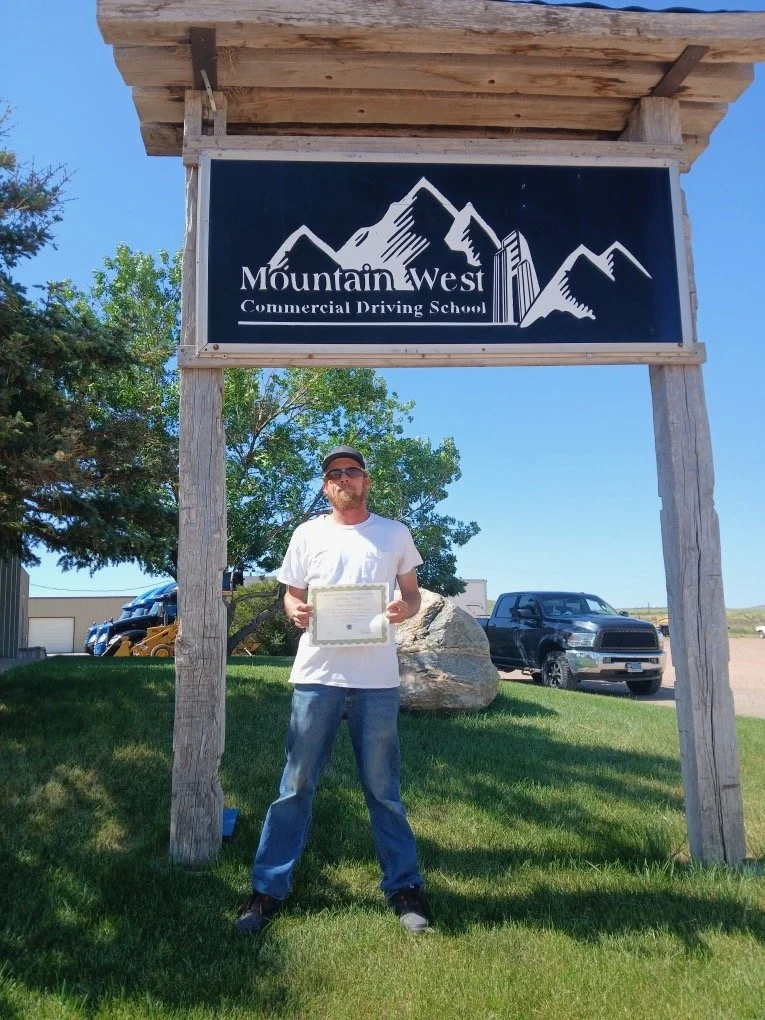 Man standing on grass holding a certificate in front of a sign that says 'Mountain West Commercial Driving School' with mountain graphics. There are trees, parked vehicles, and a blue sky in the background.