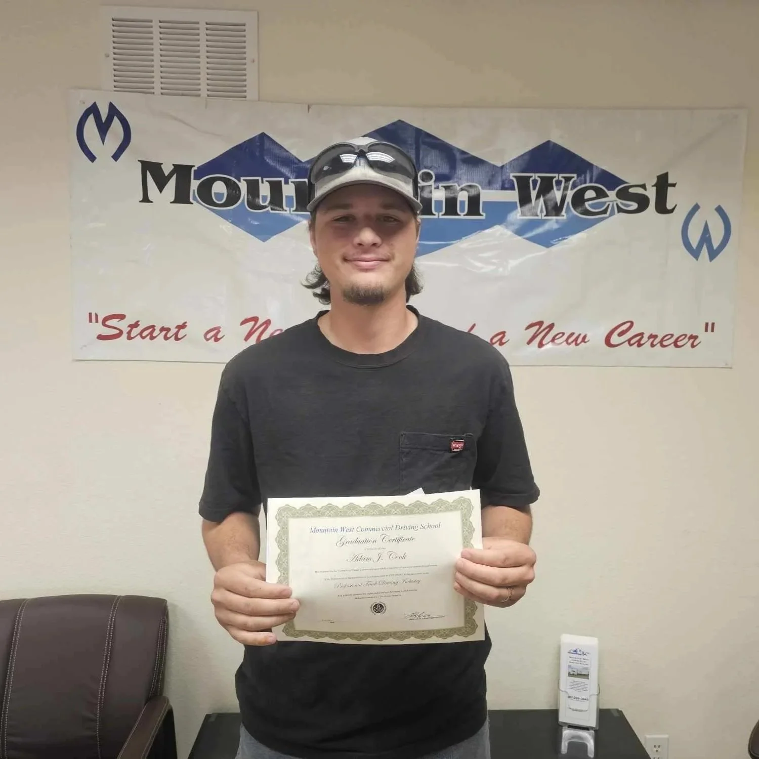 A young man holding a graduation certificate standing in front of a Mountain West banner that reads "Start a New Career."