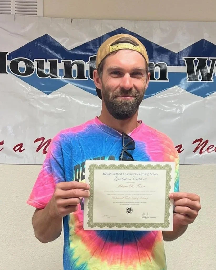 A man with a beard and mustache, wearing a yellow backwards cap and pink, blue, and yellow tie-dye shirt, holding a certificate in front of a banner that partially reads 'Mountain W...'. He is also wearing sunglasses hanging from his shirt collar.