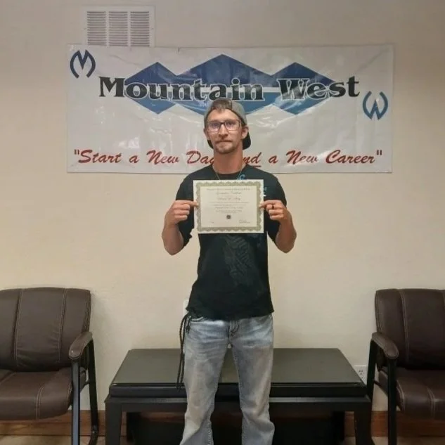 A young man with glasses holding a certificate in front of a banner that says 'Mountain West' and the slogan 'Start a New Day and a New Career', standing between two chairs in a room.