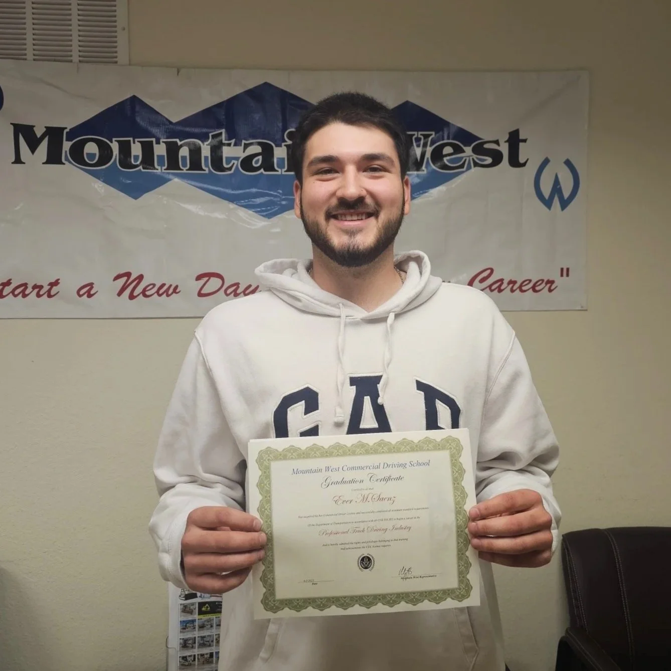 Smiling young man with dark hair and beard holding a graduation certificate, standing in front of a Mountain West banner.