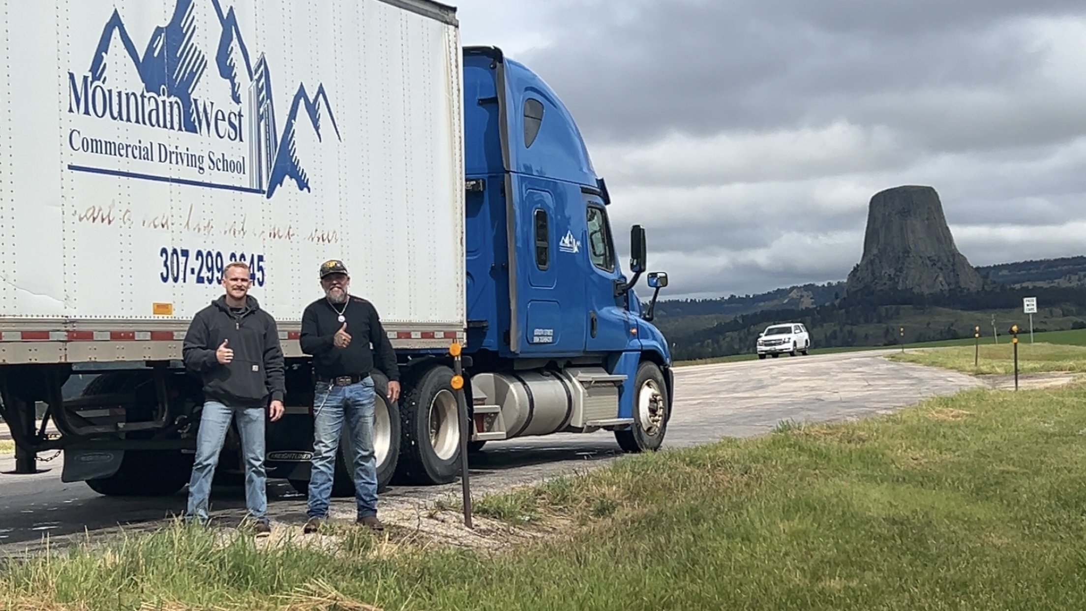 Two men standing beside a blue truck in a rural area with a large rock formation in the background. One man is giving a thumbs-up.