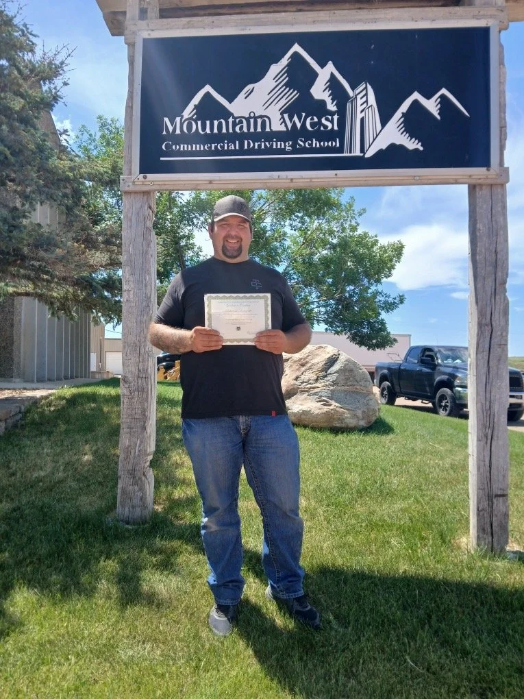 Man holding a certificate standing outdoors in front of a sign for Mountain West Commercial Driving School with mountains graphic, green grass, trees, and parked cars visible.