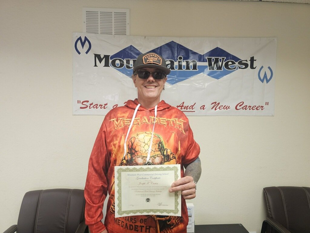 A smiling man wearing sunglasses, a black cap, and a Megadeth hoodie is holding a certificate. Behind him is a Mountain West banner on a wall.